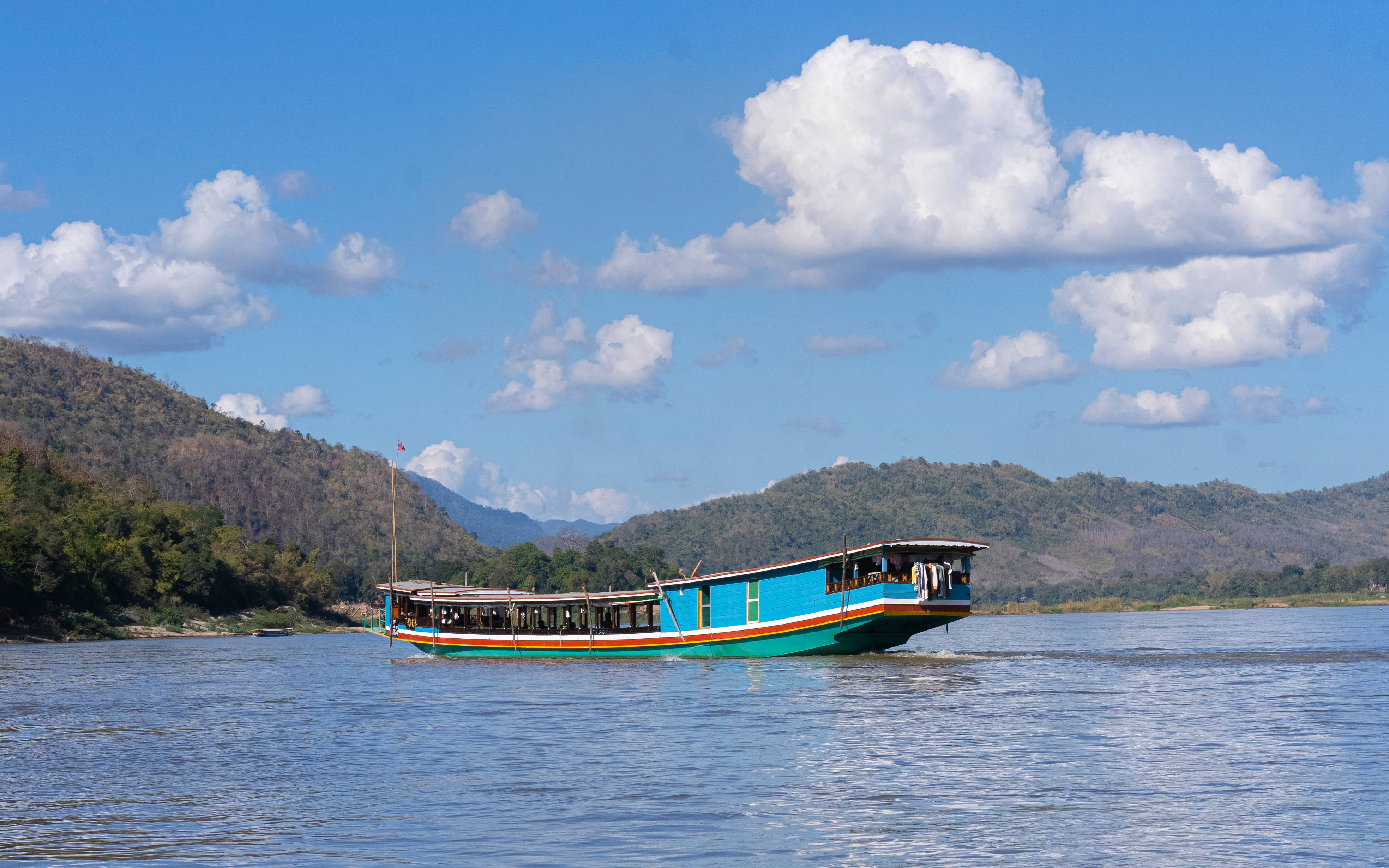 Laos 桌布 壁紙 — 一艘充滿活力的藍色長尾船在平靜的河面上滑行，頭頂是明亮的藍天，點綴著蓬鬆的白雲。