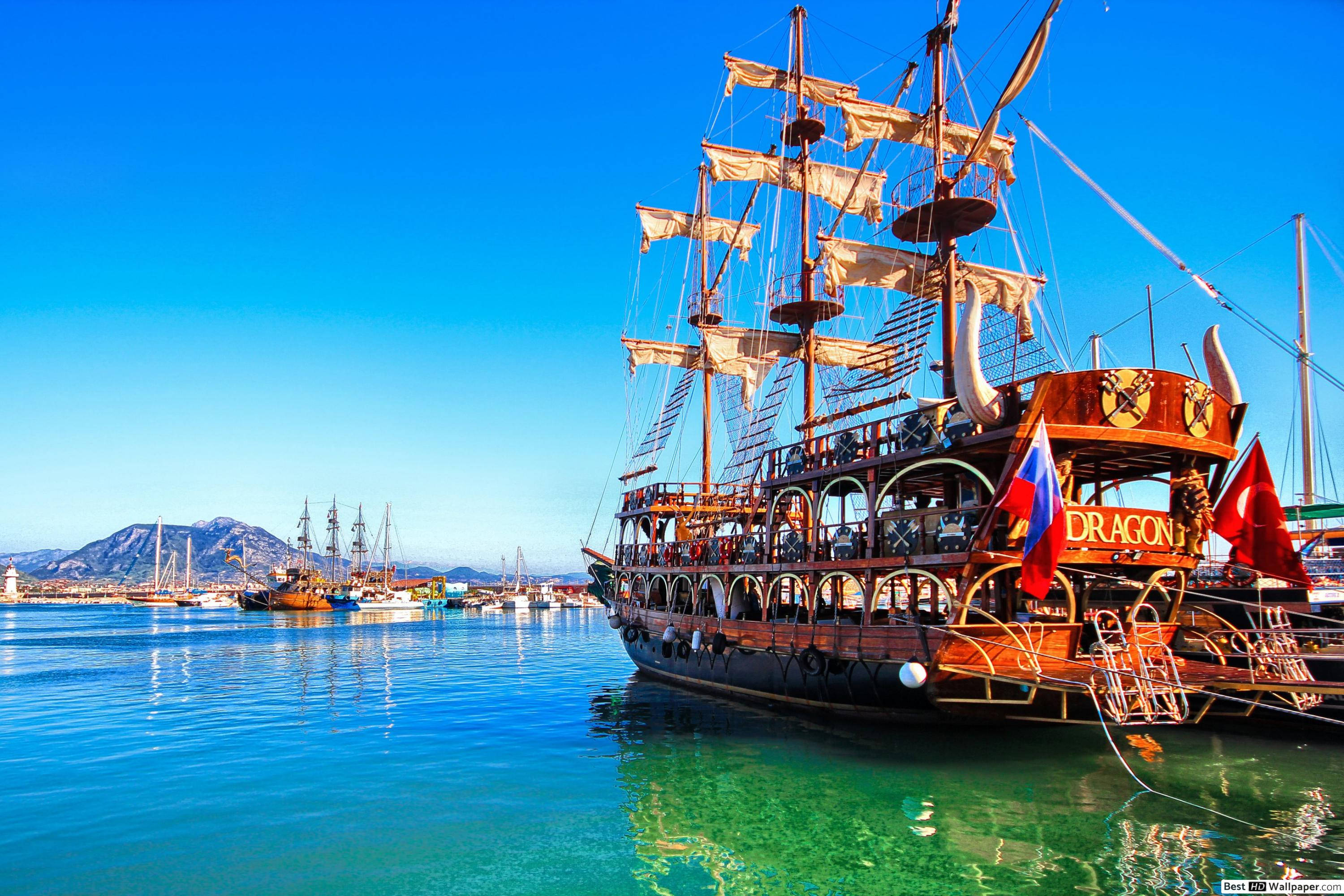 Sailing Ship 桌布 壁紙 — 一艘名為Dragon的大型木製海盜船，帆布張開，停泊在碧綠的海水中，背景是晴朗的藍天和遠山。