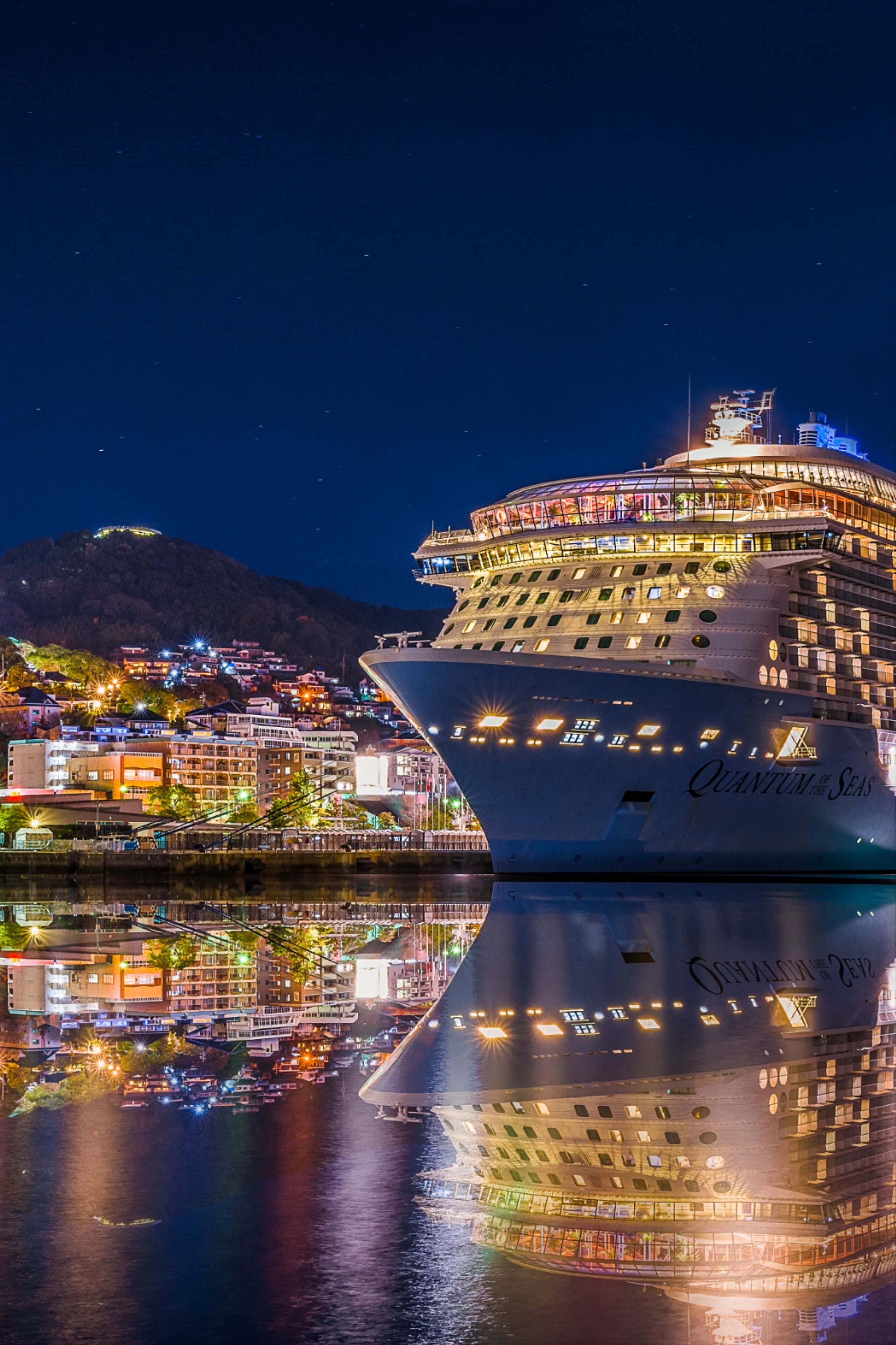 Cruise 桌布 壁紙 — 一艘名為海洋量子號的巨型郵輪在繁星點點的夜空下散發著溫暖的光芒，其倒影在平靜的水面上閃爍，旁邊是一個燈火通明的沿海小鎮。