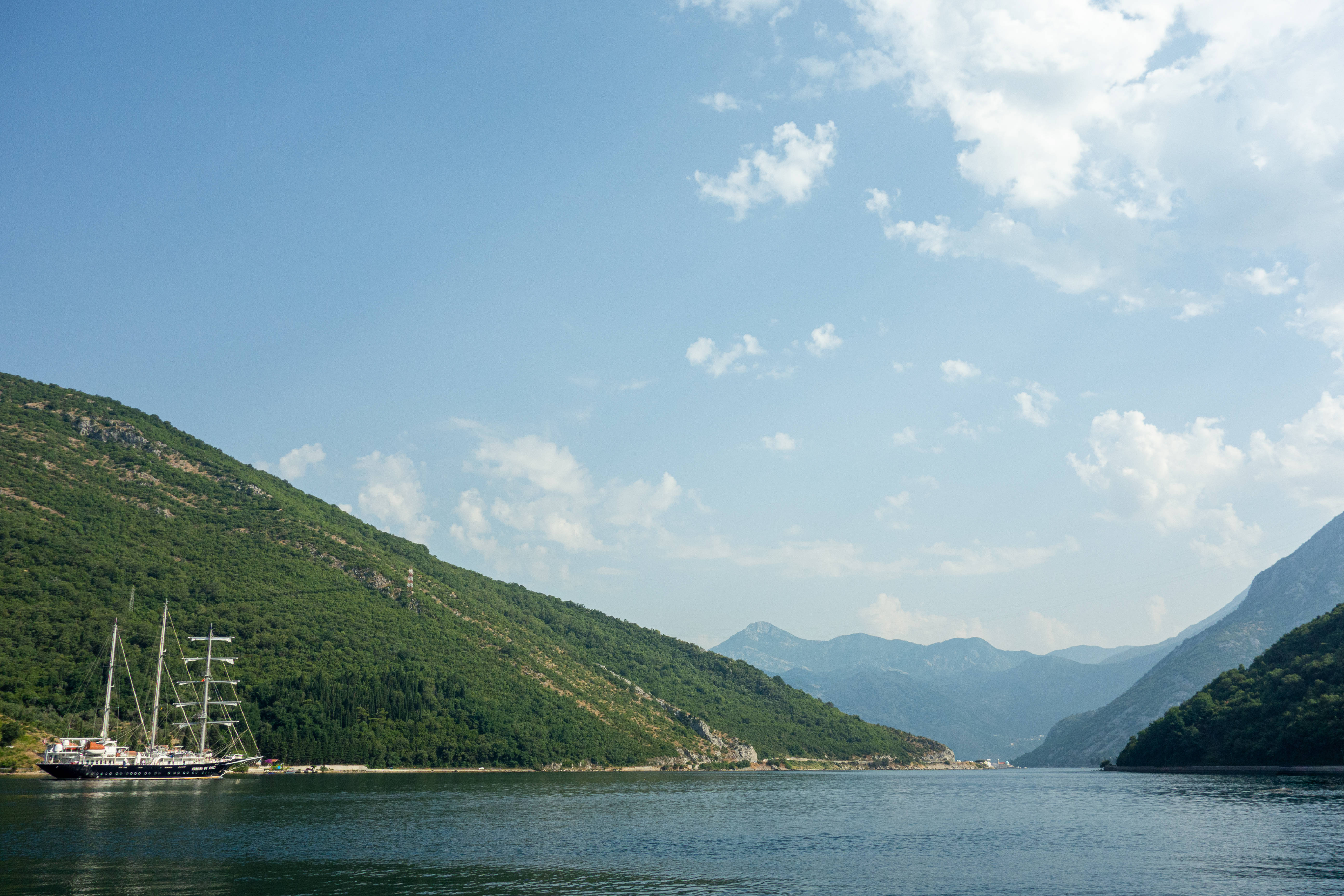 Montenegro 桌布 壁紙 — 一艘大型帆船在深藍色峽灣中寧靜地停泊，兩側是鬱鬱蔥蔥的綠色山脈，天空明亮，雲朵散佈。