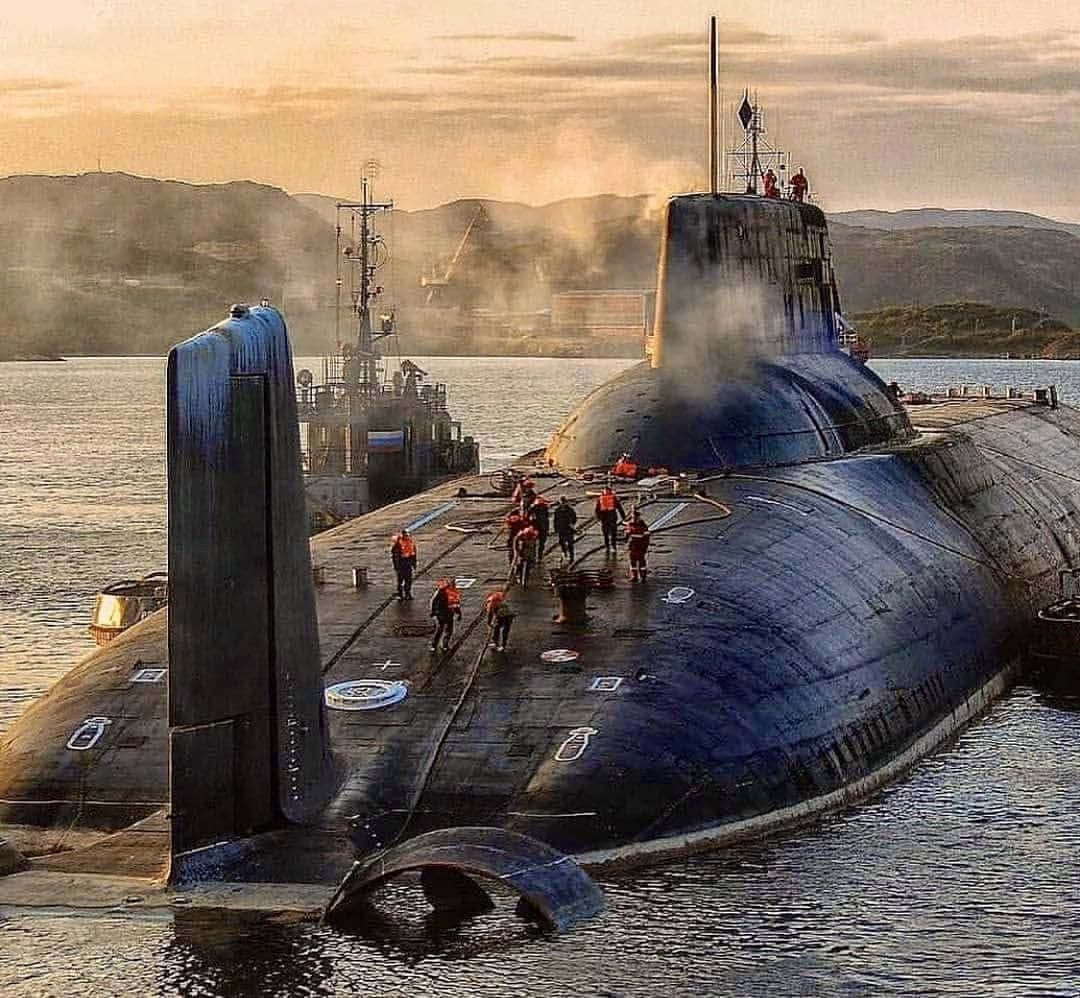 Submarine 桌布 壁紙 — 一艘大型深色潛艇停靠在碼頭，身穿橙色背心的船員在甲板上行走，蒸汽在金色日落天空的映襯下升起。