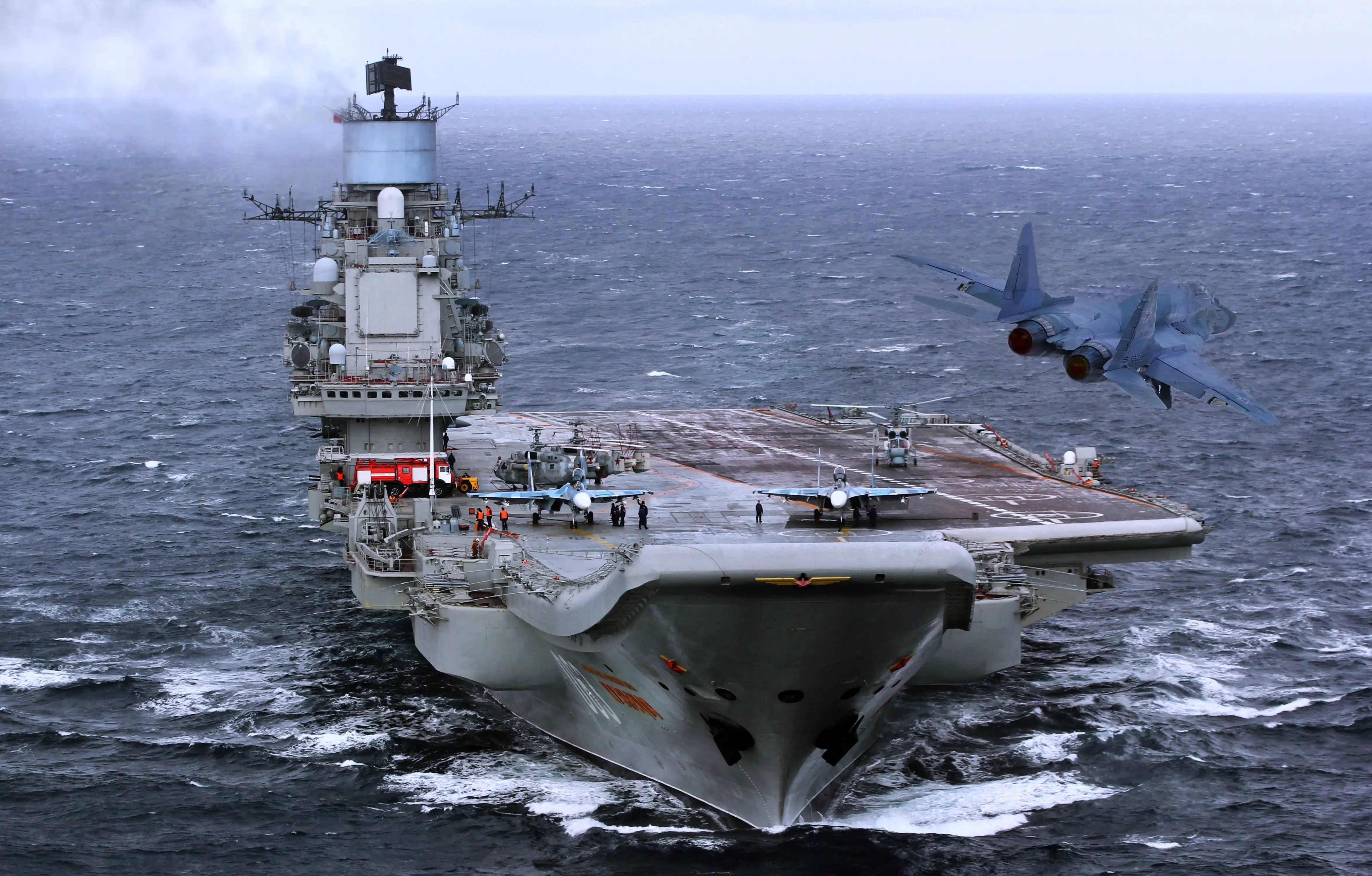 Air Force 桌布 壁紙 — 一艘大型灰色航空母艦在波濤洶湧的深藍色海洋中航行，戰鬥機正在起飛，其他戰機則停放在甲板上。