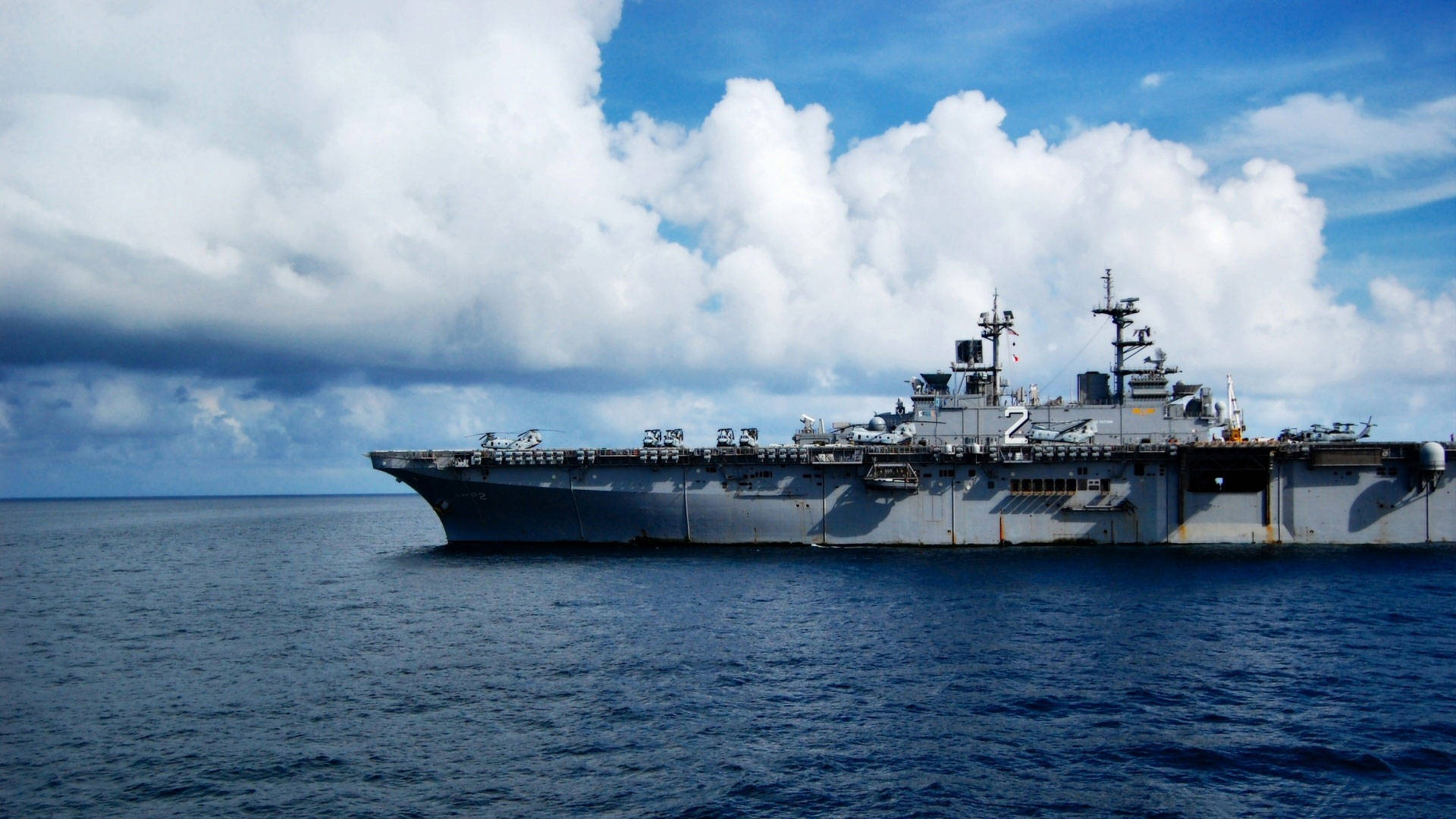 Warship 桌布 壁紙 — 一艘大型灰色航空母艦在深藍色海洋上平靜航行，天空佈滿蓬鬆的白雲，景象壯麗。