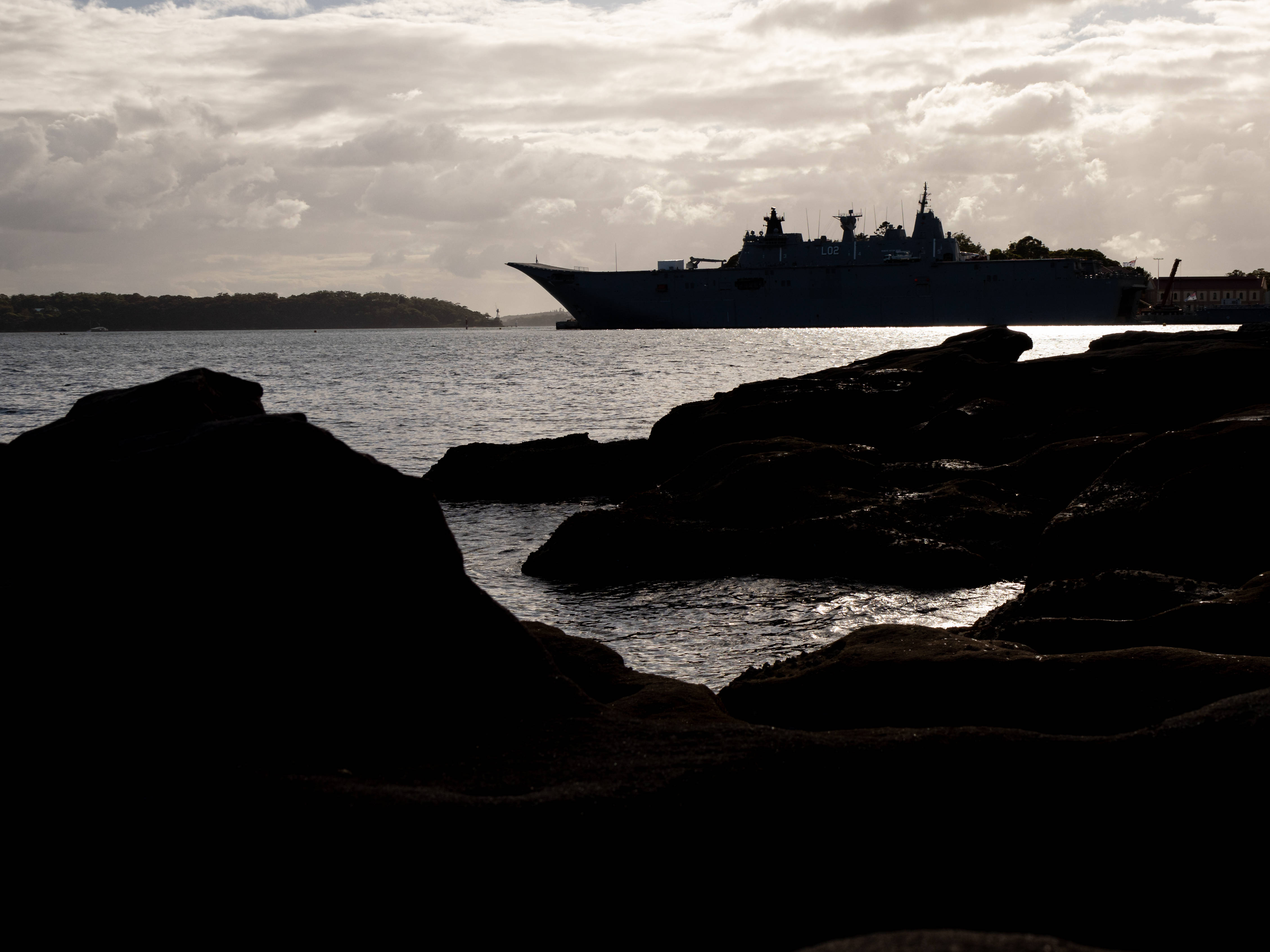 Warship 桌布 壁紙 — 一艘大型軍艦在陰雲密佈的天空下呈現剪影，靜止在平靜的水面上，前景有黑暗的岩石框住。