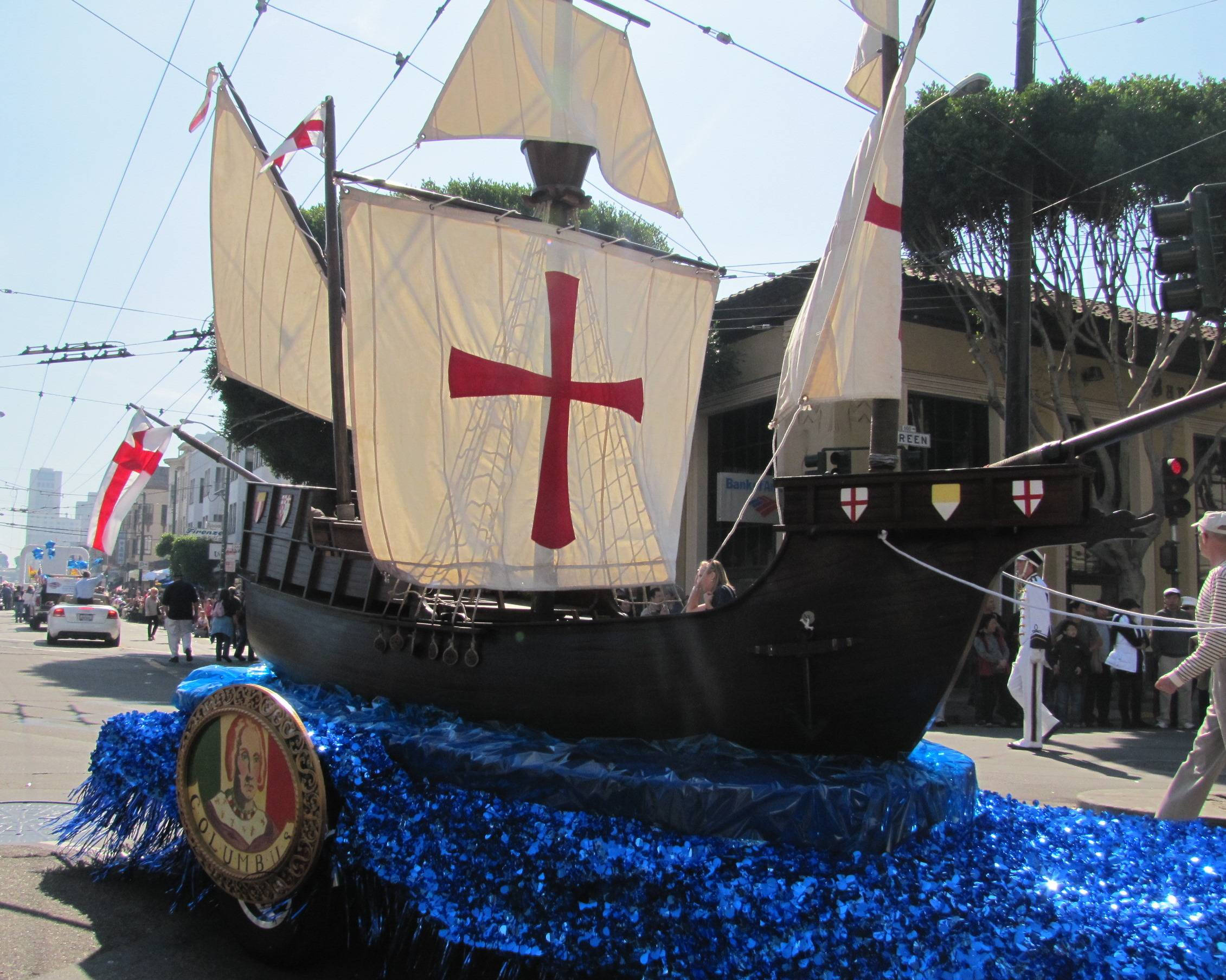 Columbus Day 桌布 壁紙 — 一艘大型黑色海盜船花車，帶有紅色十字帆和藍色閃光波浪，在陽光明媚的城市街道上巡遊。
