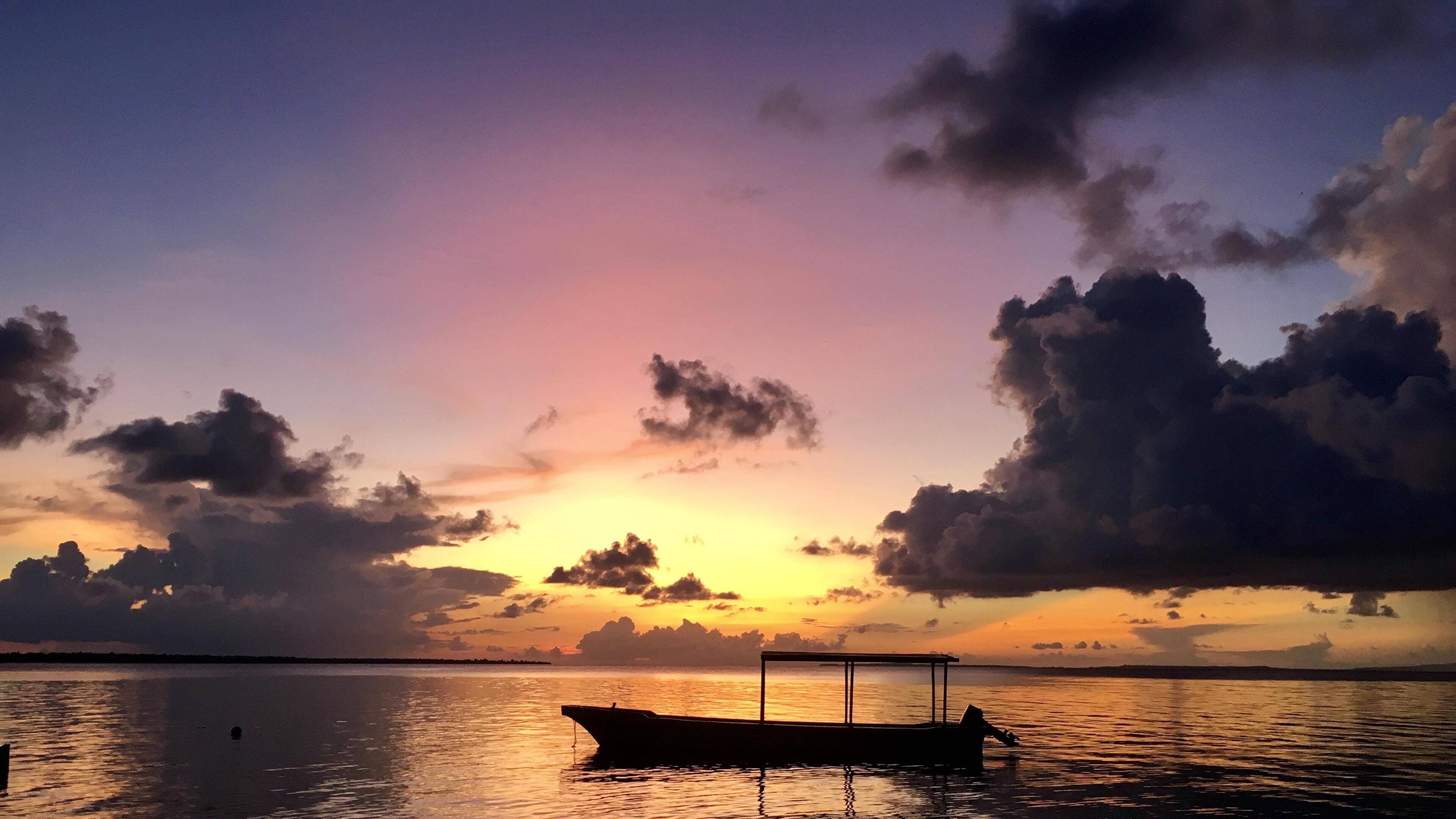 Palau 桌布 壁紙 — 一艘孤獨的小船在充滿活力的日落天空下形成剪影，紫色、橙色和黃色的色調映照在平靜的水面上。