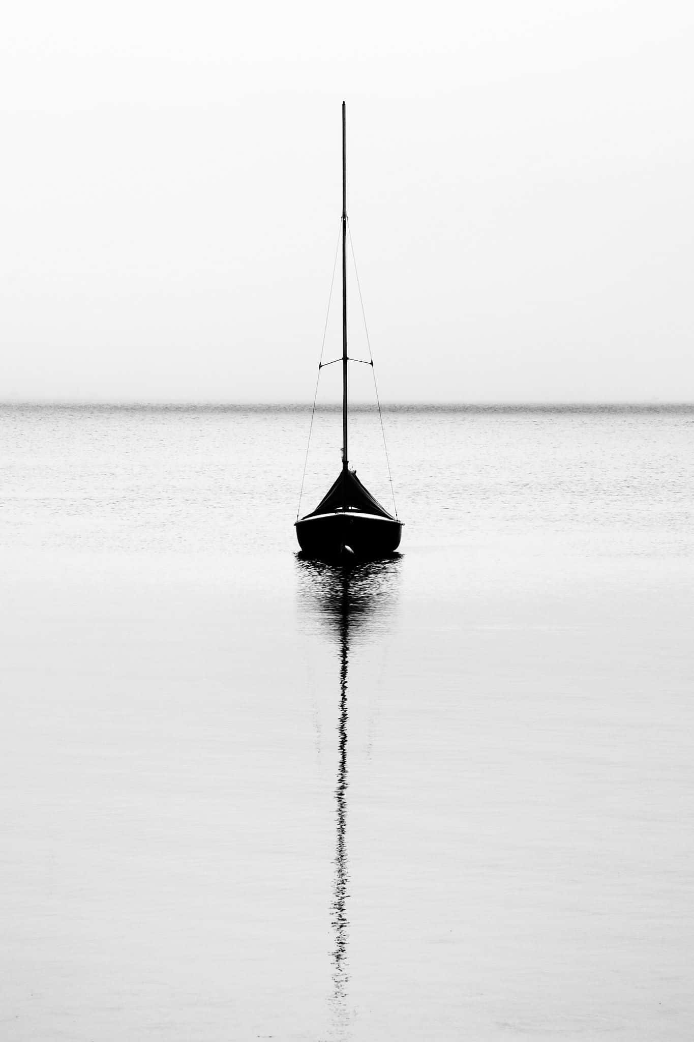 Minimalist Photography 桌布 壁紙 — 一艘孤獨的帆船剪影在平靜的水面上寧靜地漂浮，在陰天之下形成完美的垂直倒影。