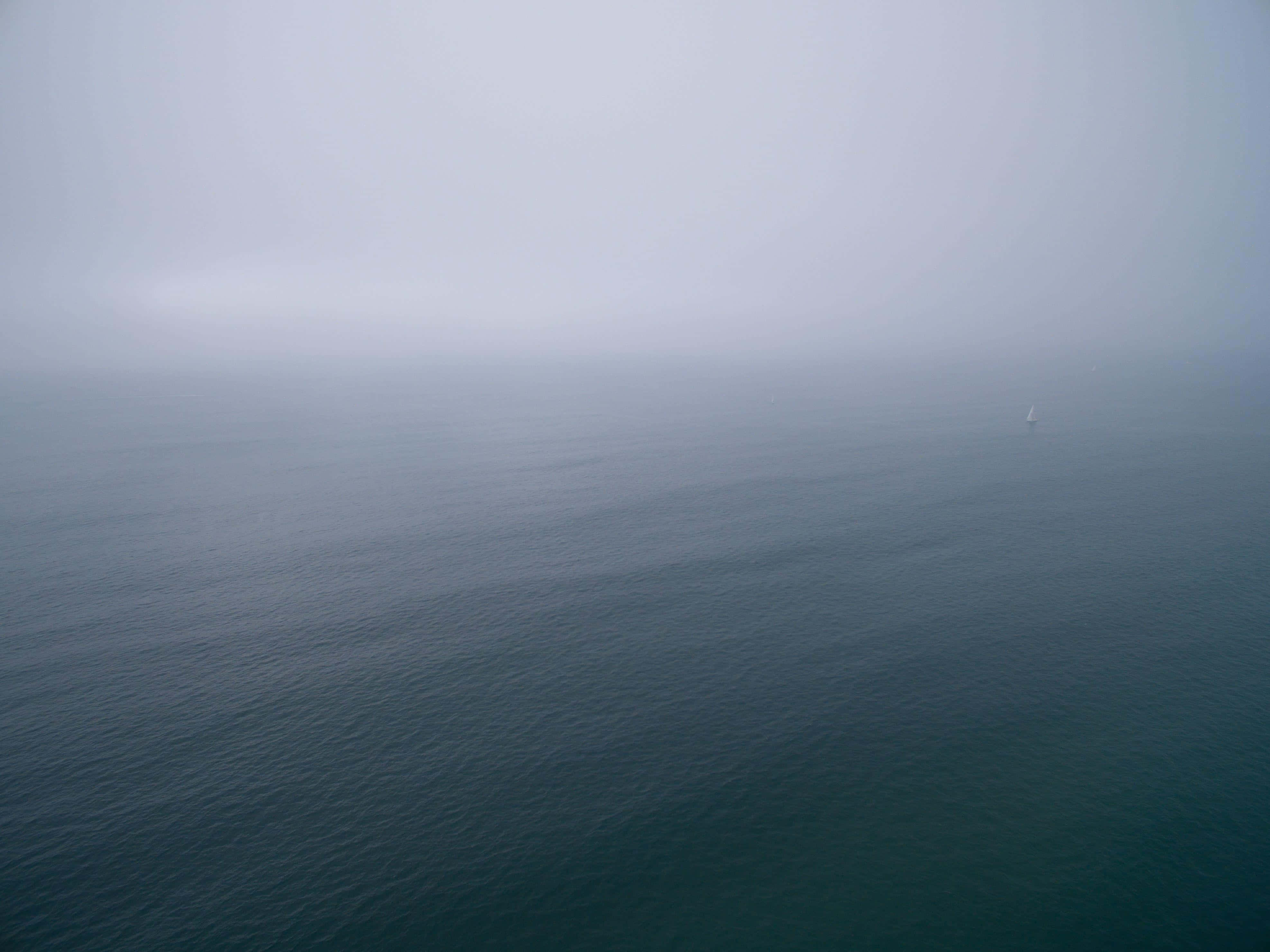 Foggy Aesthetic 桌布 壁紙 — 一艘孤獨的帆船在灰色天空下，在廣闊平靜的海洋上逐漸消失在霧中。
