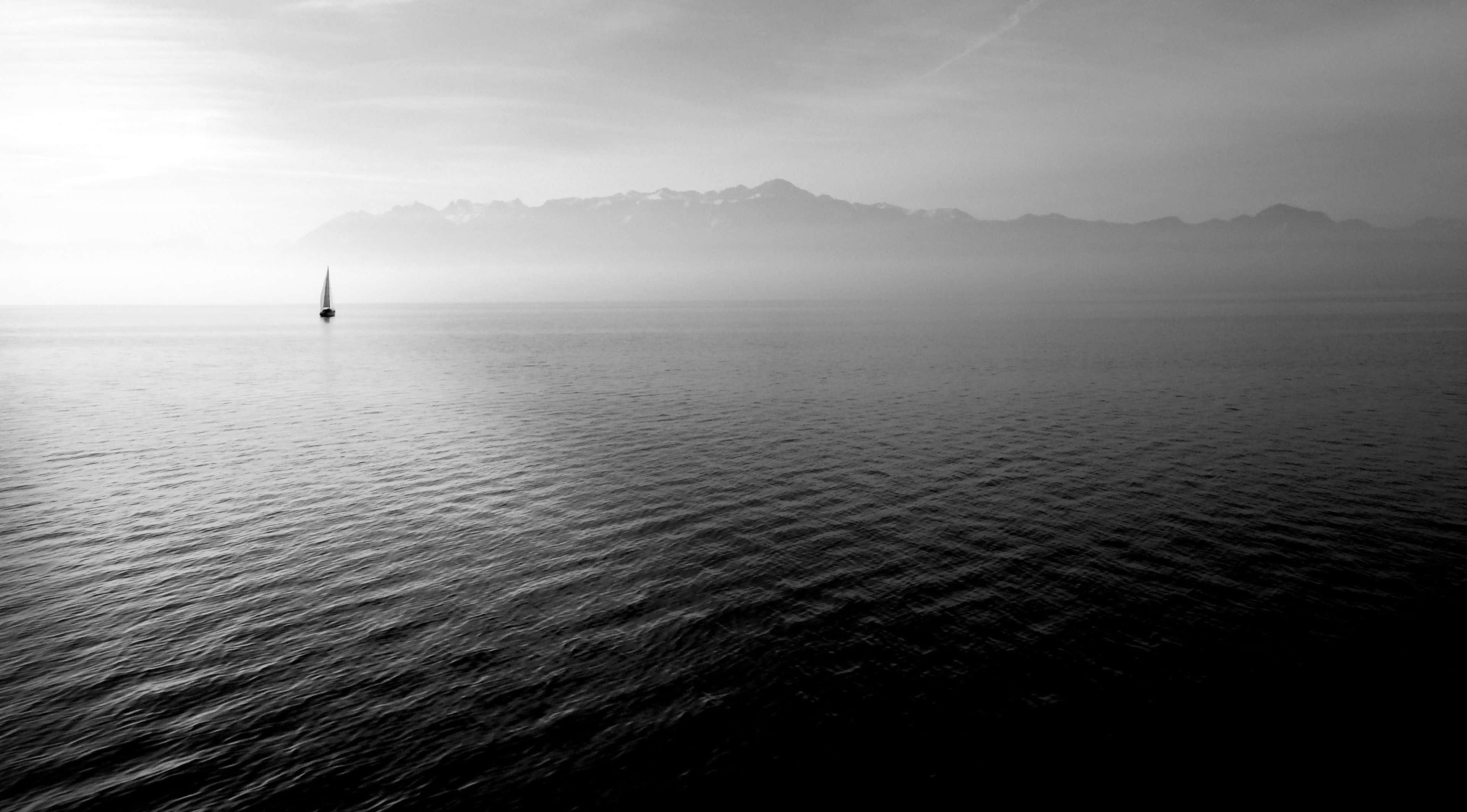 Black And White Ocean 桌布 壁紙 — 一艘孤獨的帆船在霧氣瀰漫的單色湖面上滑行，遠處是白雪覆蓋的山脈，天空是柔和的黎明。