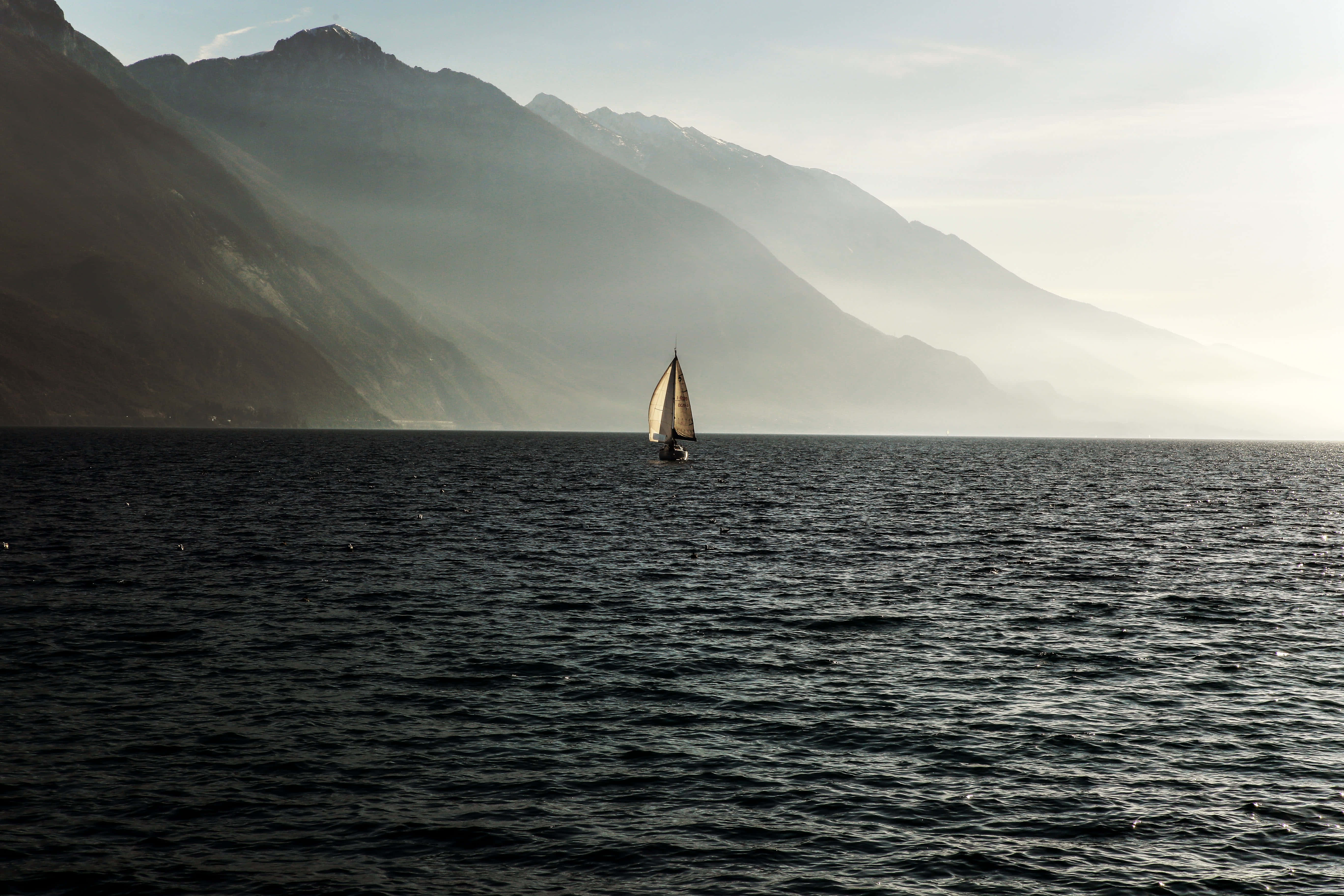 Sailboat 桌布 壁紙 — 一艘孤獨的帆船在霧氣瀰漫的湖面上滑行，高聳的山脈在柔和的黎明天空下籠罩在薄霧中。