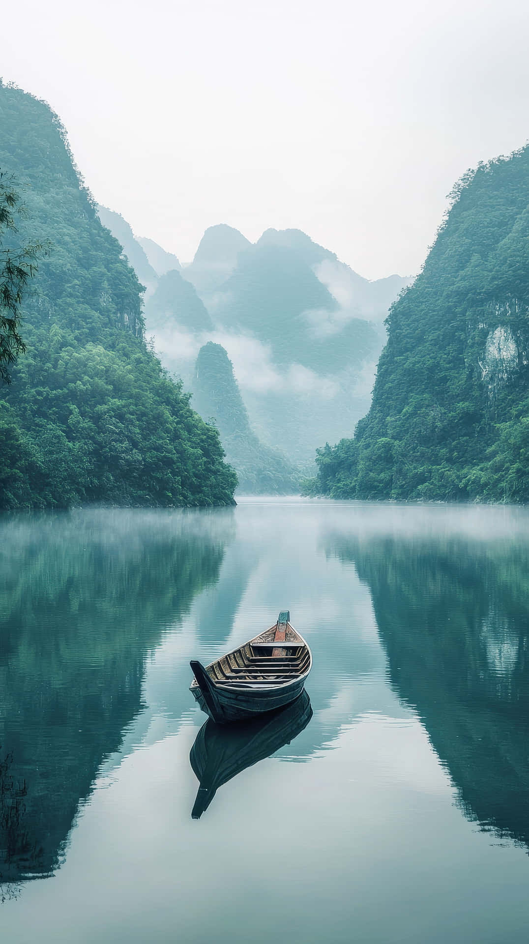 Serene wooden boat floating on a misty lake surrounded by lush mountains 壁紙 — 一艘孤獨的木船在霧氣瀰漫的湖面上靜靜漂浮，四周是高聳的綠色山脈，山頂上籠罩著薄霧。