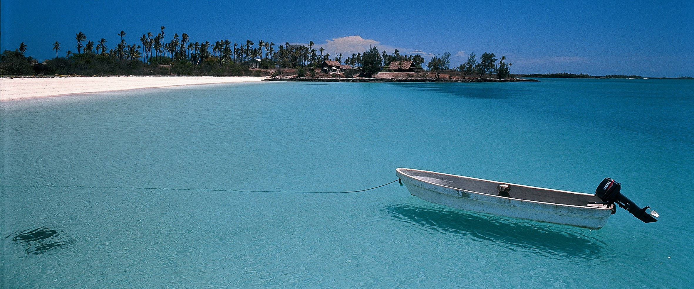 Mozambique 桌布 壁紙 — 一艘孤獨的白色機動船漂浮在清澈碧綠的海水中，靠近一條 pristine 白色沙灘，沙灘上排列著棕櫚樹和質樸的小屋，上方是明亮的藍天。