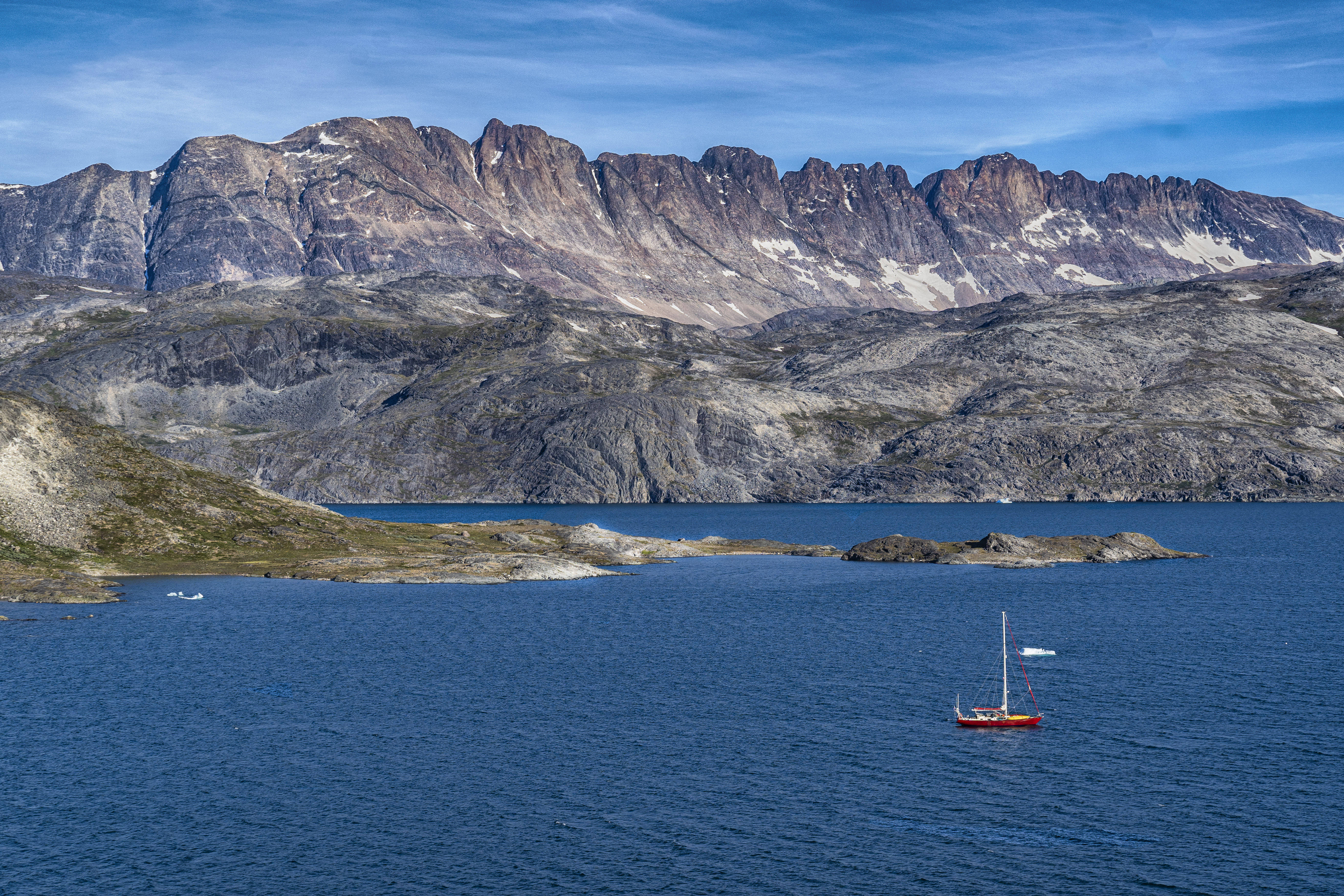 Greenland 桌布 壁紙 — 一艘孤獨的紅色帆船在深藍色的水面上滑行，下方是崎嶇的岩石山脈，上方是清澈的藍天。