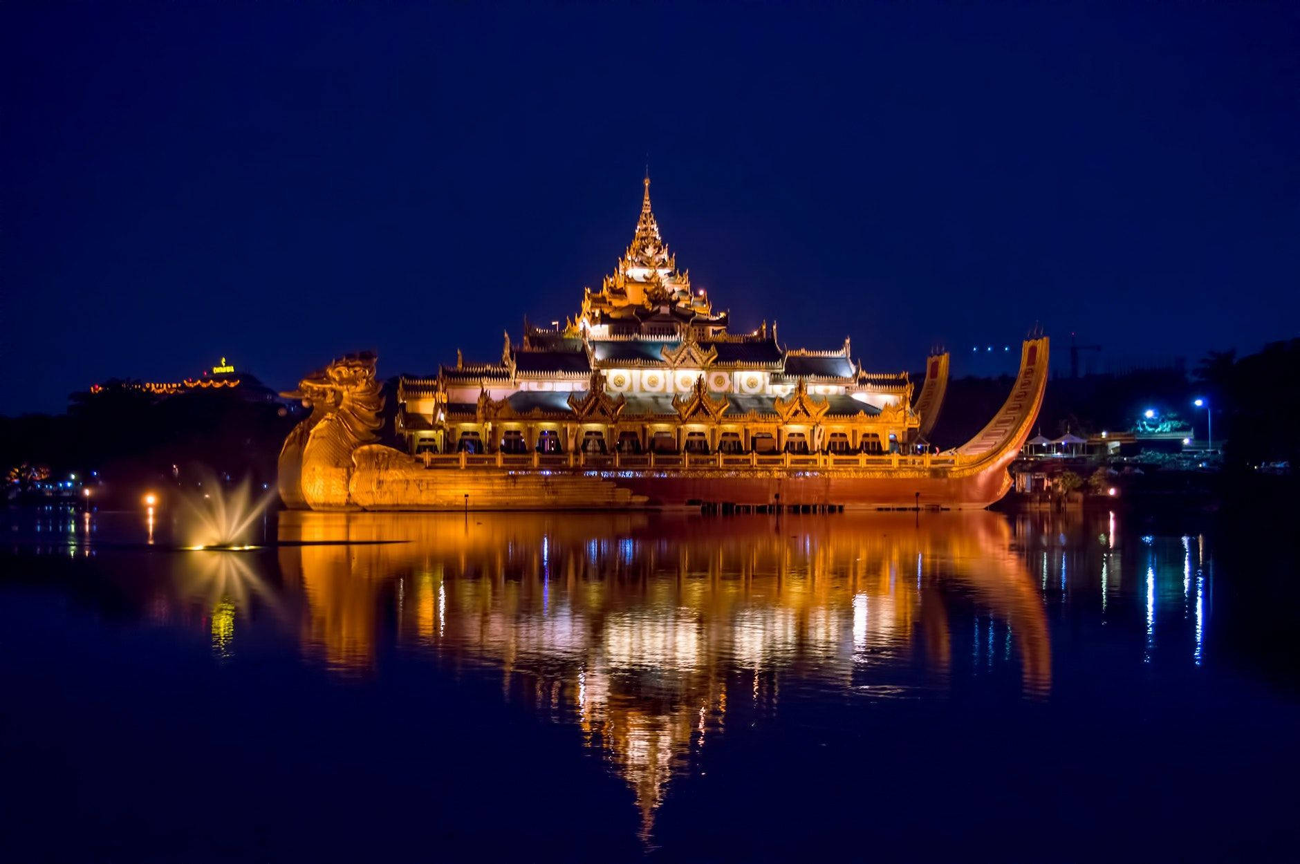 Yangon 桌布 壁紙 — 一艘宏偉的燈光寺廟船在夜晚平靜地漂浮在黑暗的水面上，金色的燈光美麗地倒映在平靜的水面中。