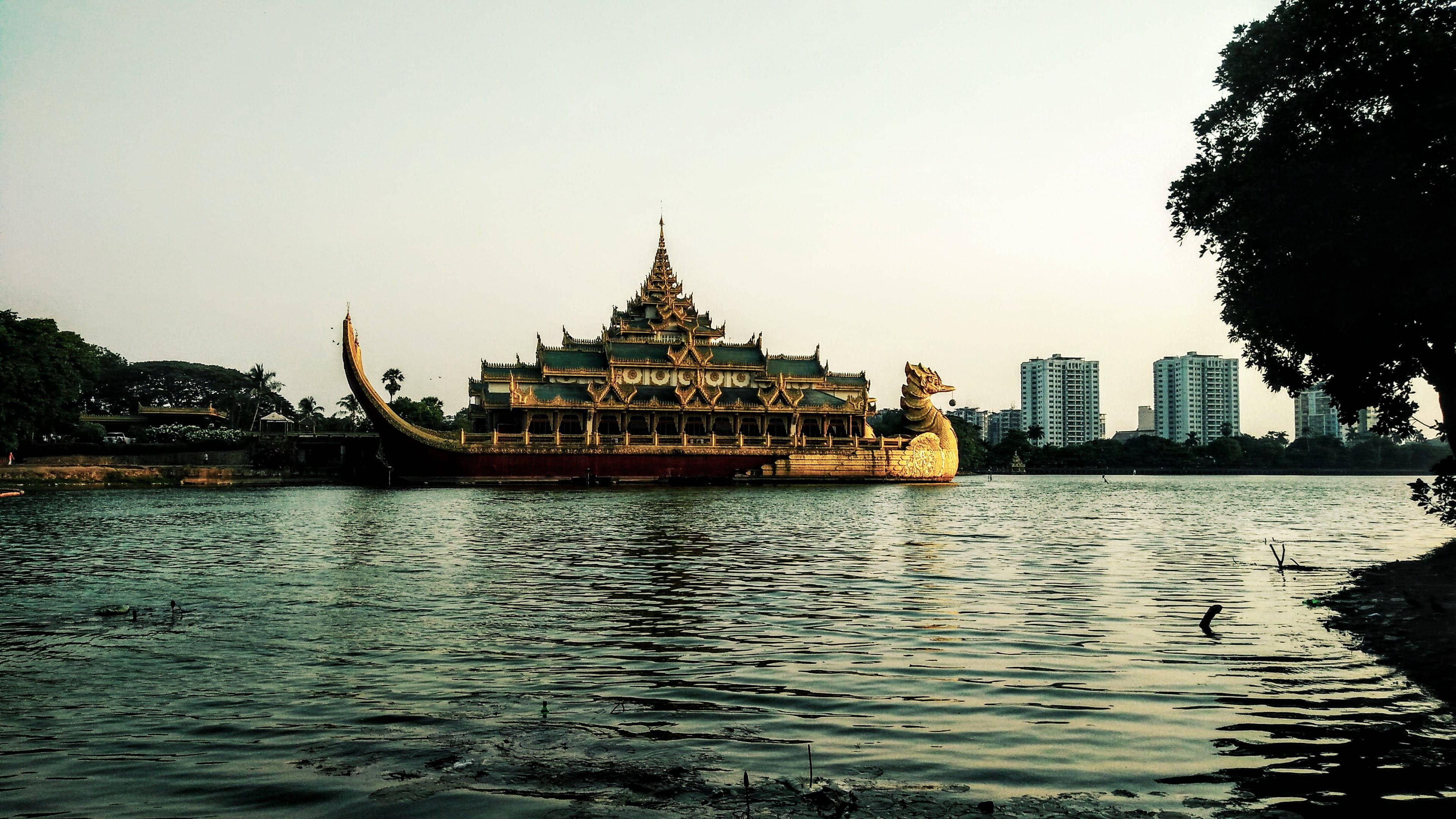 Yangon 桌布 壁紙 — 一艘宏偉的金色寺廟船在黃昏時平靜地漂浮在平靜的水面上，背景是現代建築和樹木。