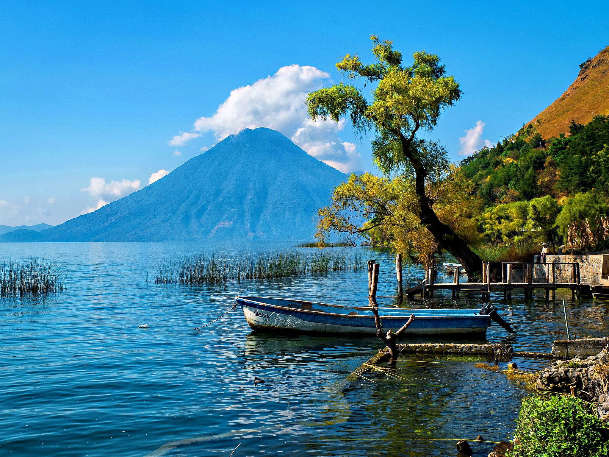 Guatemala 桌布 壁紙 — 一艘寧靜的藍色小船停靠在木製碼頭旁，旁邊是寧靜的湖泊，雄偉的火山在晴朗的天空下聳立。