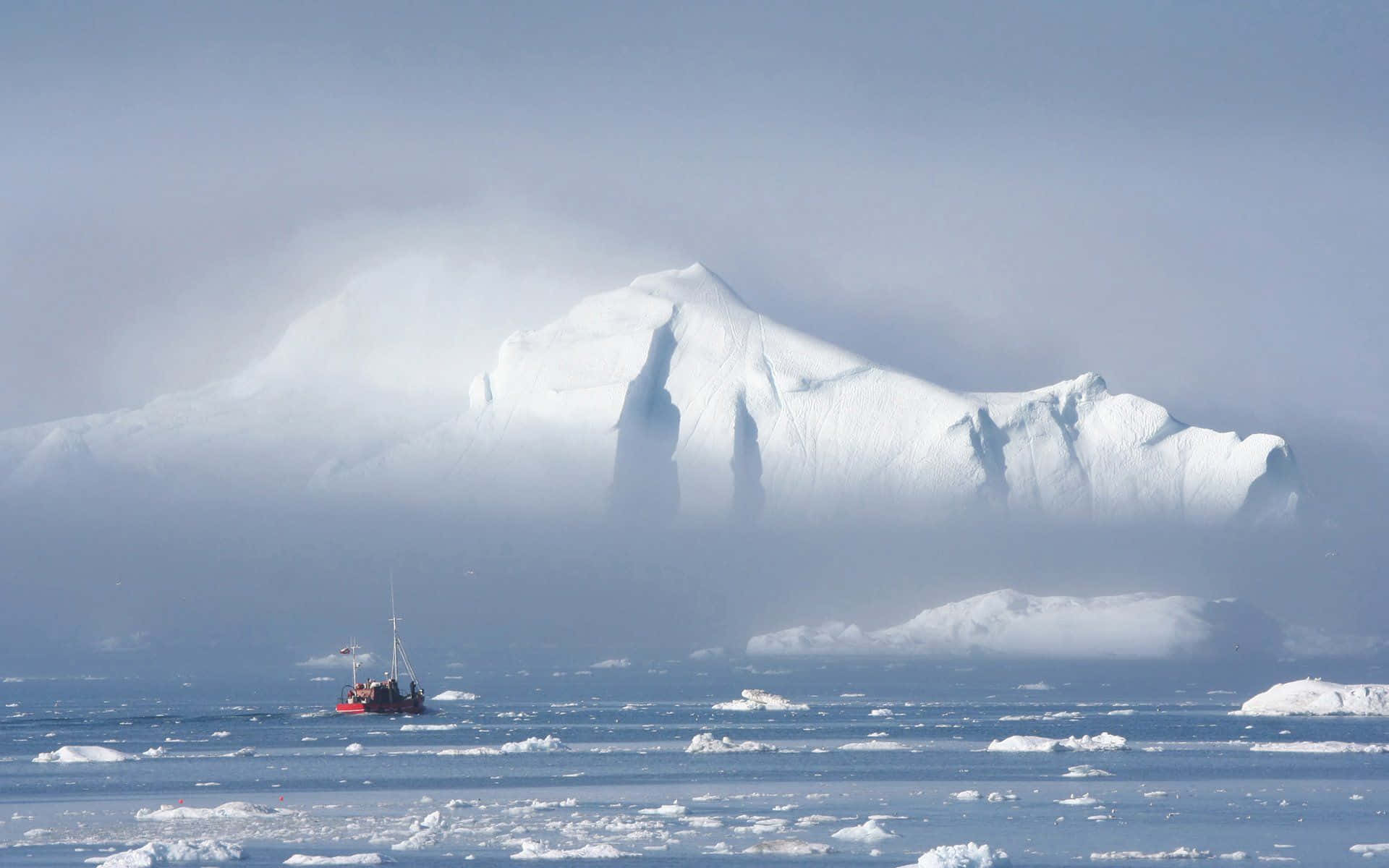 Greenland 桌布 壁紙 — 一艘小紅船在嚴酷的極地景觀中，於霧氣籠罩的巨大冰山下的冰冷水域中航行。