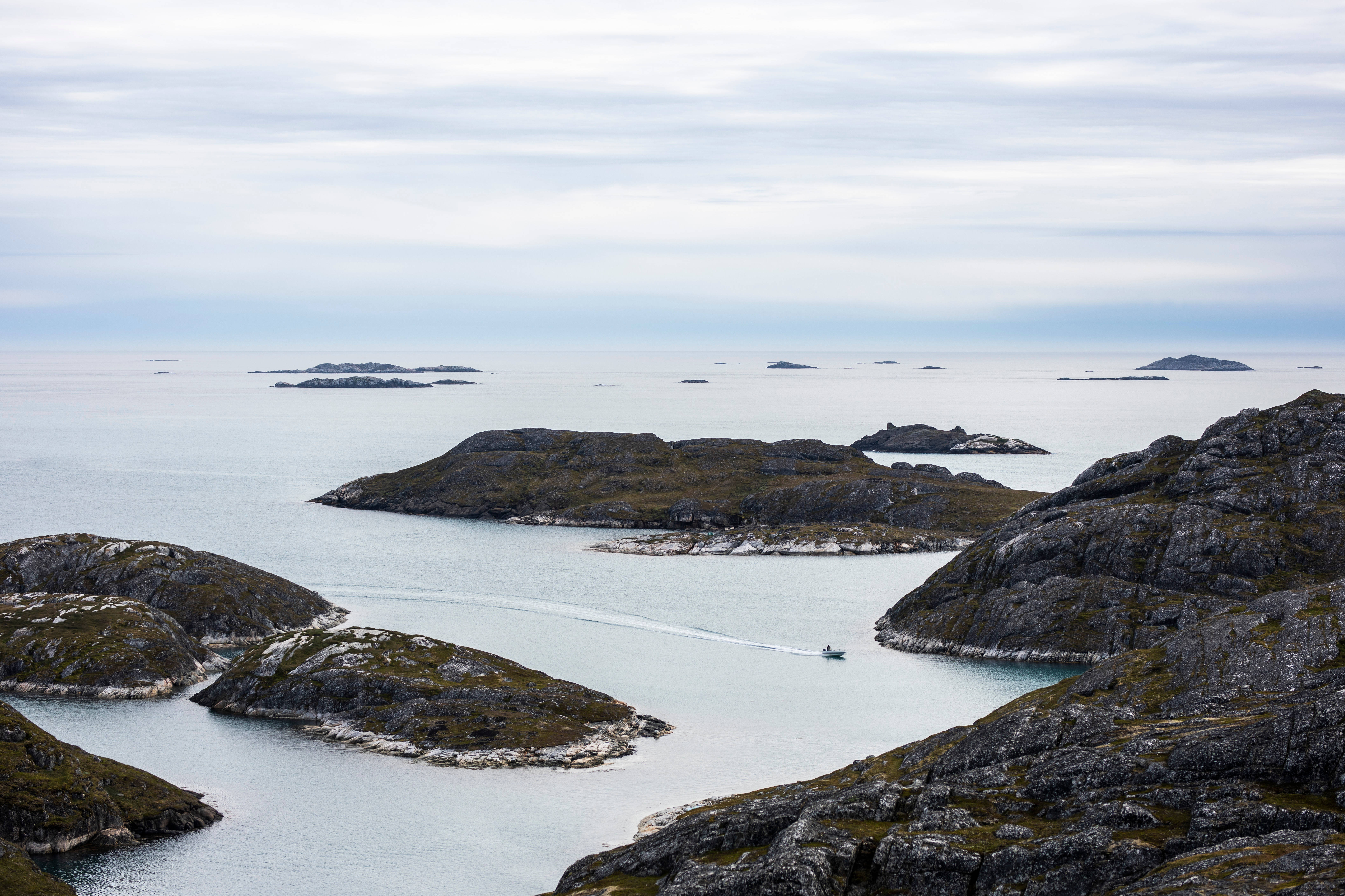 Greenland 桌布 壁紙 — 一艘小船在陰天之下，於崎嶇岩石島嶼之間的平靜水域中穿行。