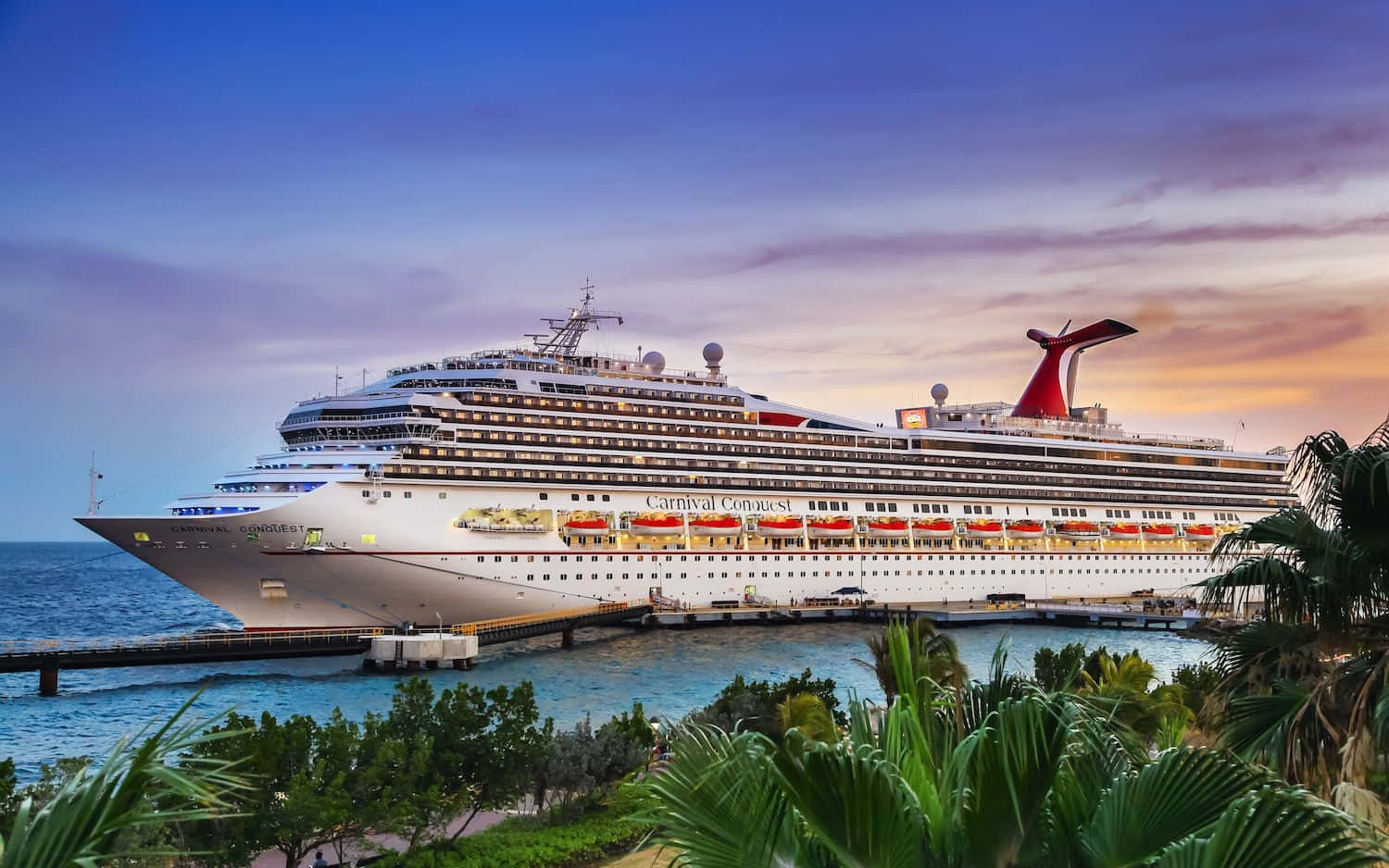 Carnival Dream 桌布 壁紙 — 一艘巨大的嘉年華征服號郵輪在黃昏時停泊，棕櫚葉框住前景，背景是充滿活力的紫色和橙色天空。