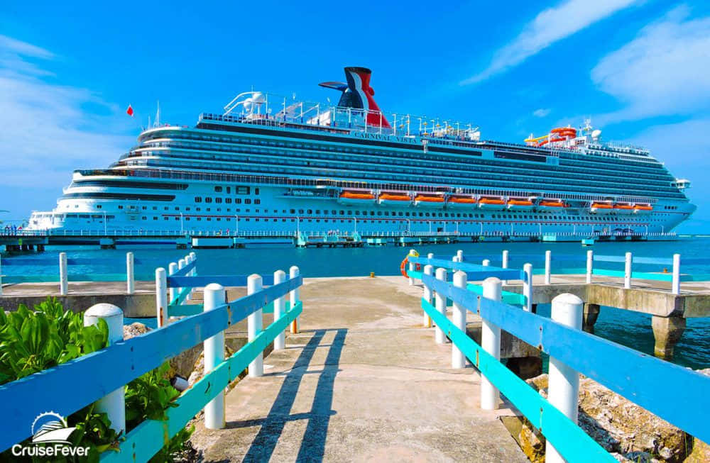 Cruise 桌布 壁紙 — 一艘巨大的嘉年華郵輪在明亮的藍天下停泊，周圍是碧綠的海水和色彩繽紛的碼頭。