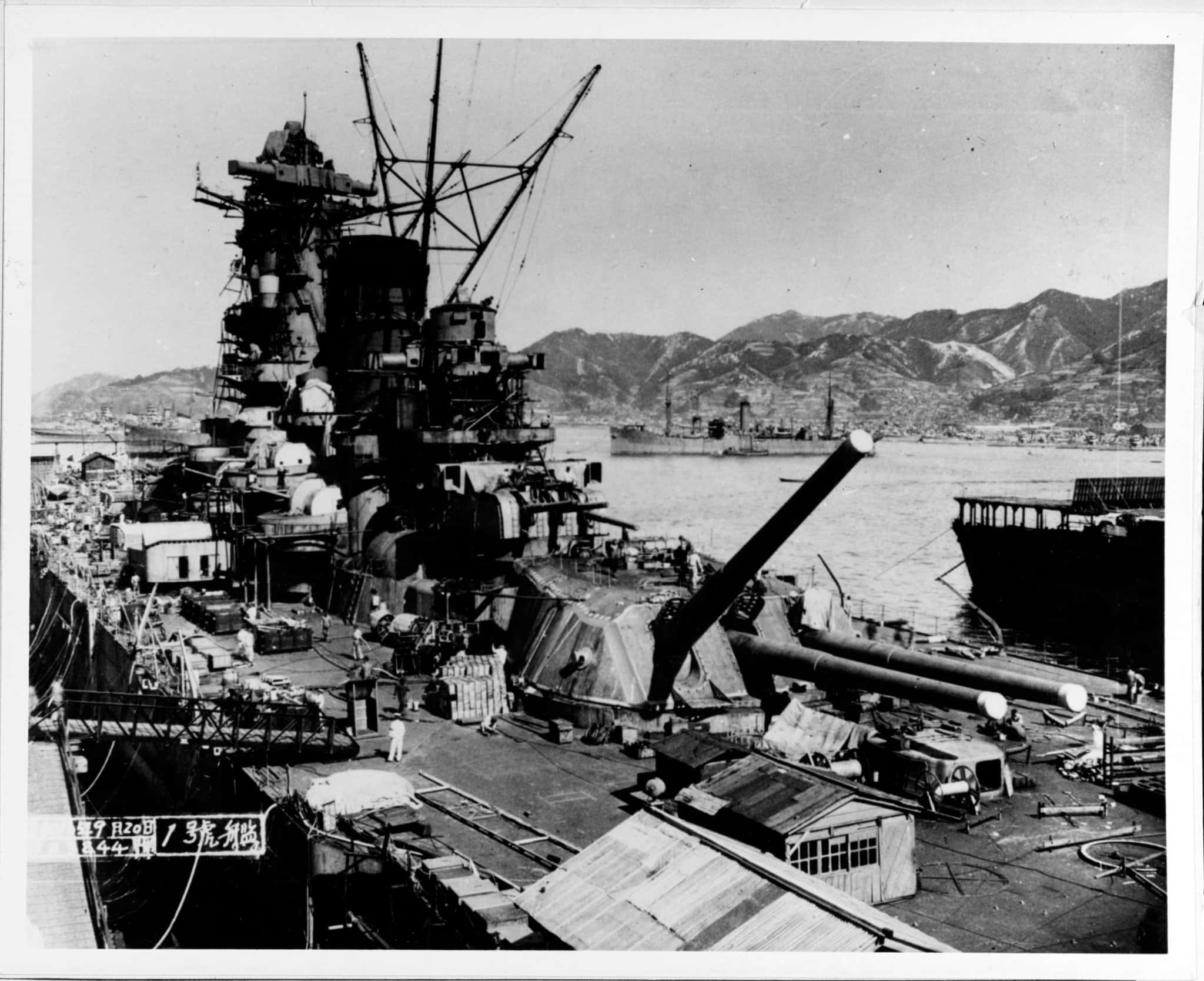 Battleships 桌布 壁紙 — 一艘巨大的戰艦主宰著港口，背景有山脈，甲板上可見船員。