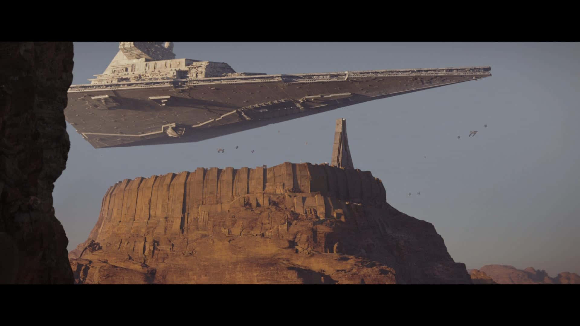 Jedha 桌布 壁紙 — 一艘巨大的星際毀滅者號盤旋在沙漠峽谷上空，峽谷中有一座高聳的巨石柱，天空朦朧。