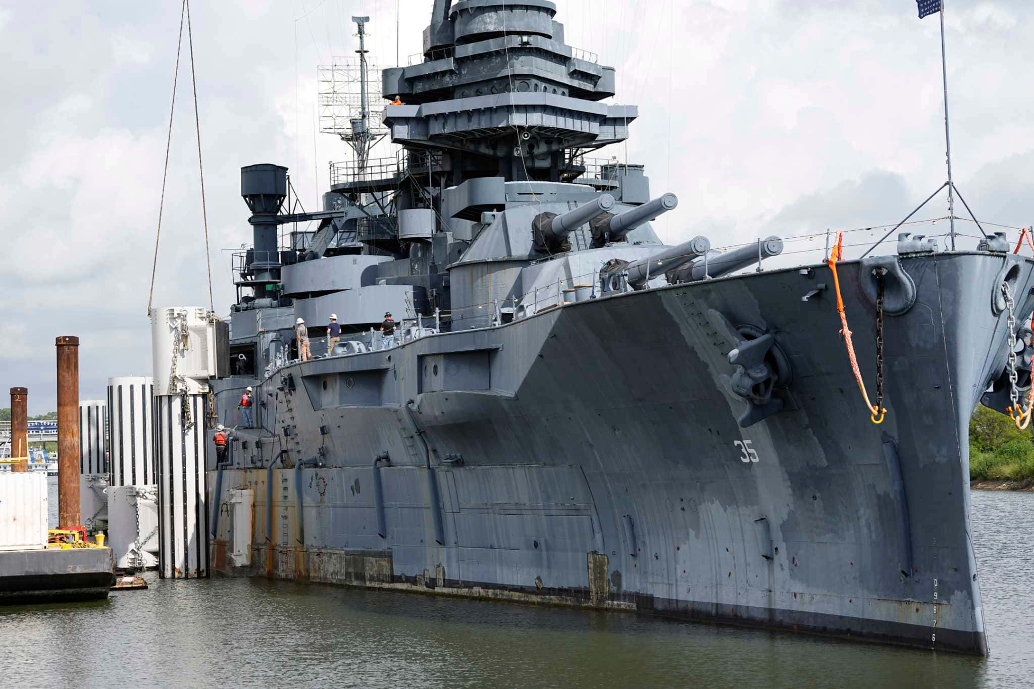 Battleships 桌布 壁紙 — 一艘巨大的灰色戰艦，帶有突出的砲塔和甲板砲，停泊在碼頭旁的平靜水域，天空多雲。
