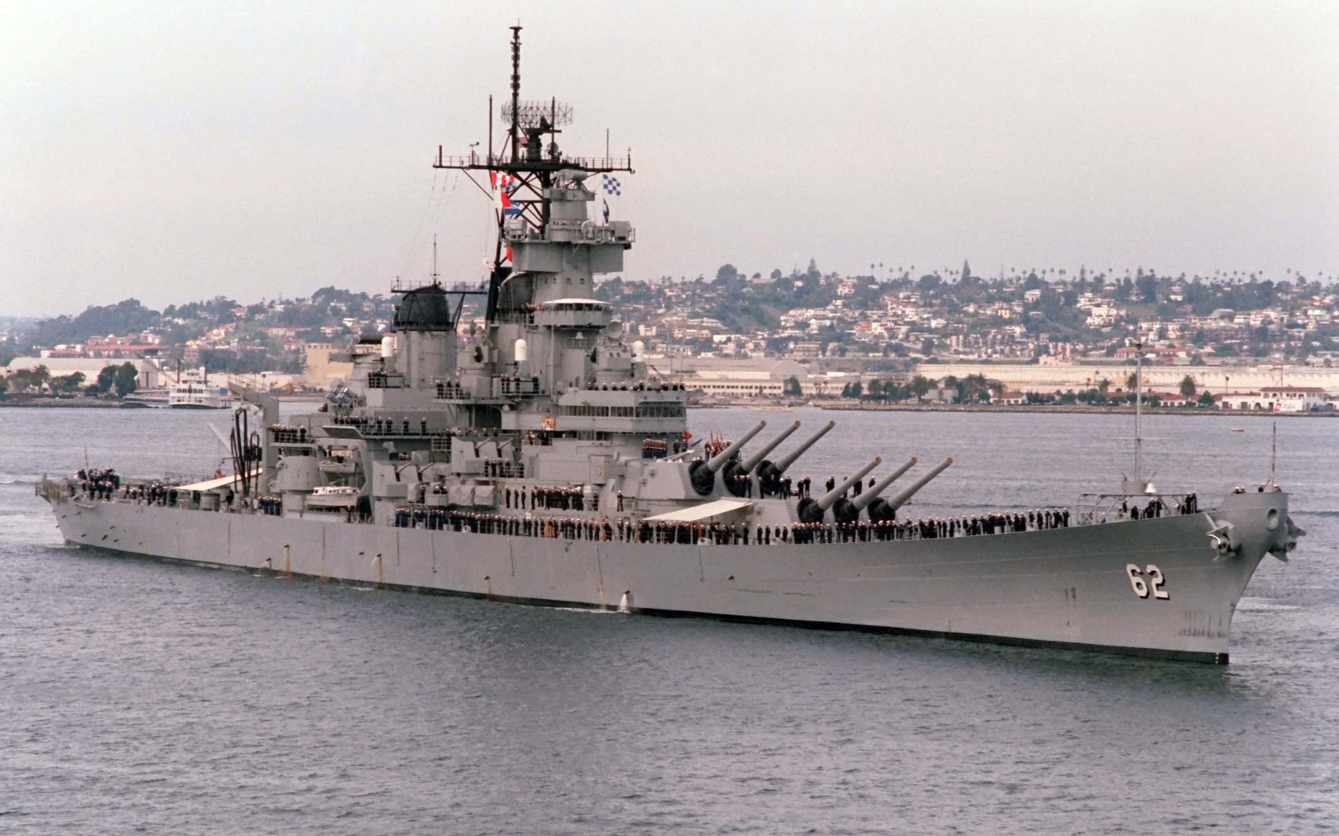 Battleships 桌布 壁紙 — 一艘巨大的灰色戰艦，編號62，在朦朧的沿海城市背景下平靜地航行。
