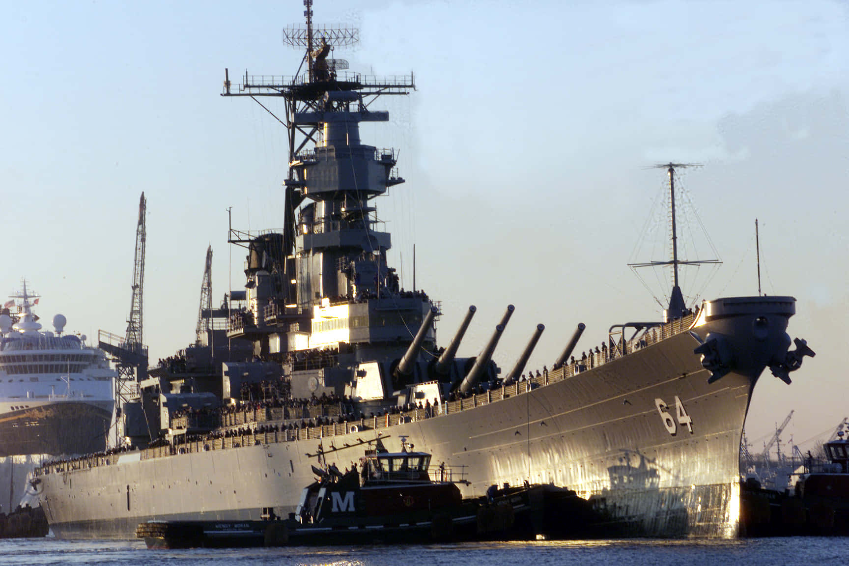 Battleships 桌布 壁紙 — 一艘巨大的灰色戰艦，編號64，帶有突出的炮塔，停泊在港口，附近有遊輪和拖船。