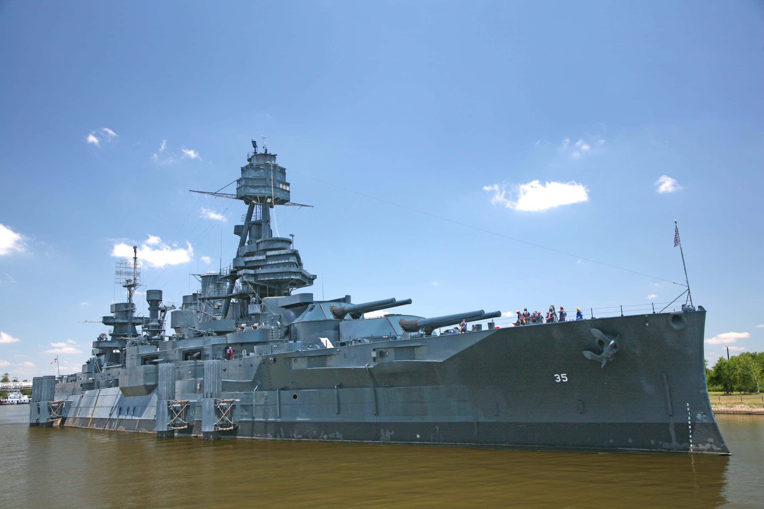 Battleships 桌布 壁紙 — 一艘巨大的灰色戰艦，配備大型火炮和高大的上層建築，漂浮在平靜的棕色水面上，天空湛藍，雲朵稀疏。