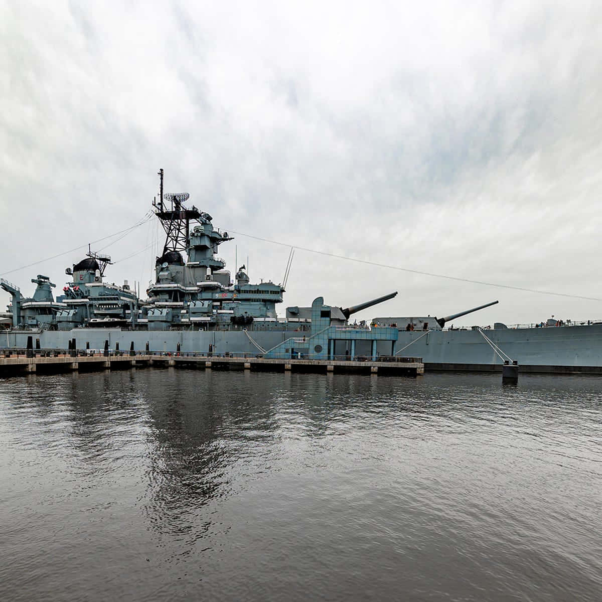 Battleships 桌布 壁紙 — 一艘巨大的灰色戰艦停泊在碼頭，天空多雲，平靜的水面映照出其宏偉的結構。