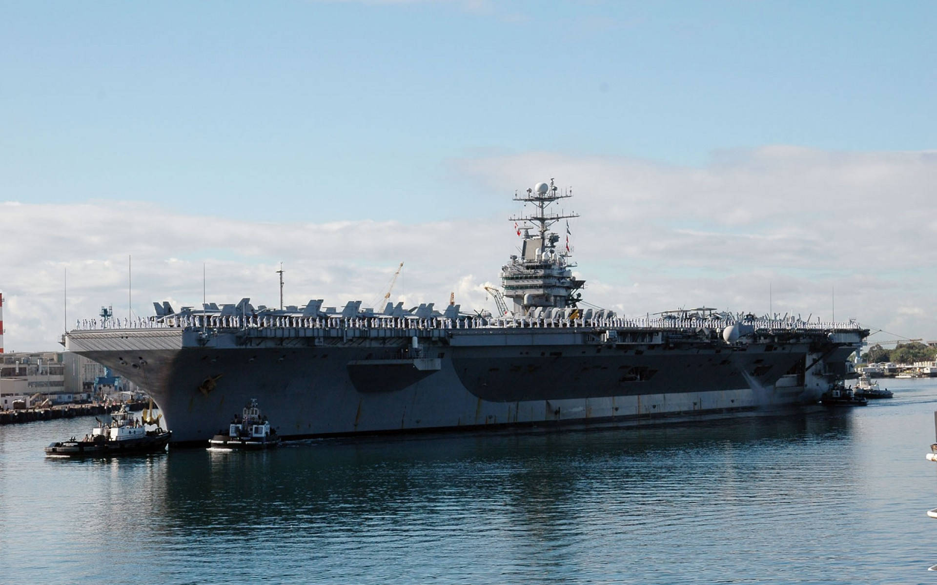 Warship 桌布 壁紙 — 一艘巨大的灰色航空母艦停泊在平靜的港灣水域中，附近有小拖船，天空部分多雲。