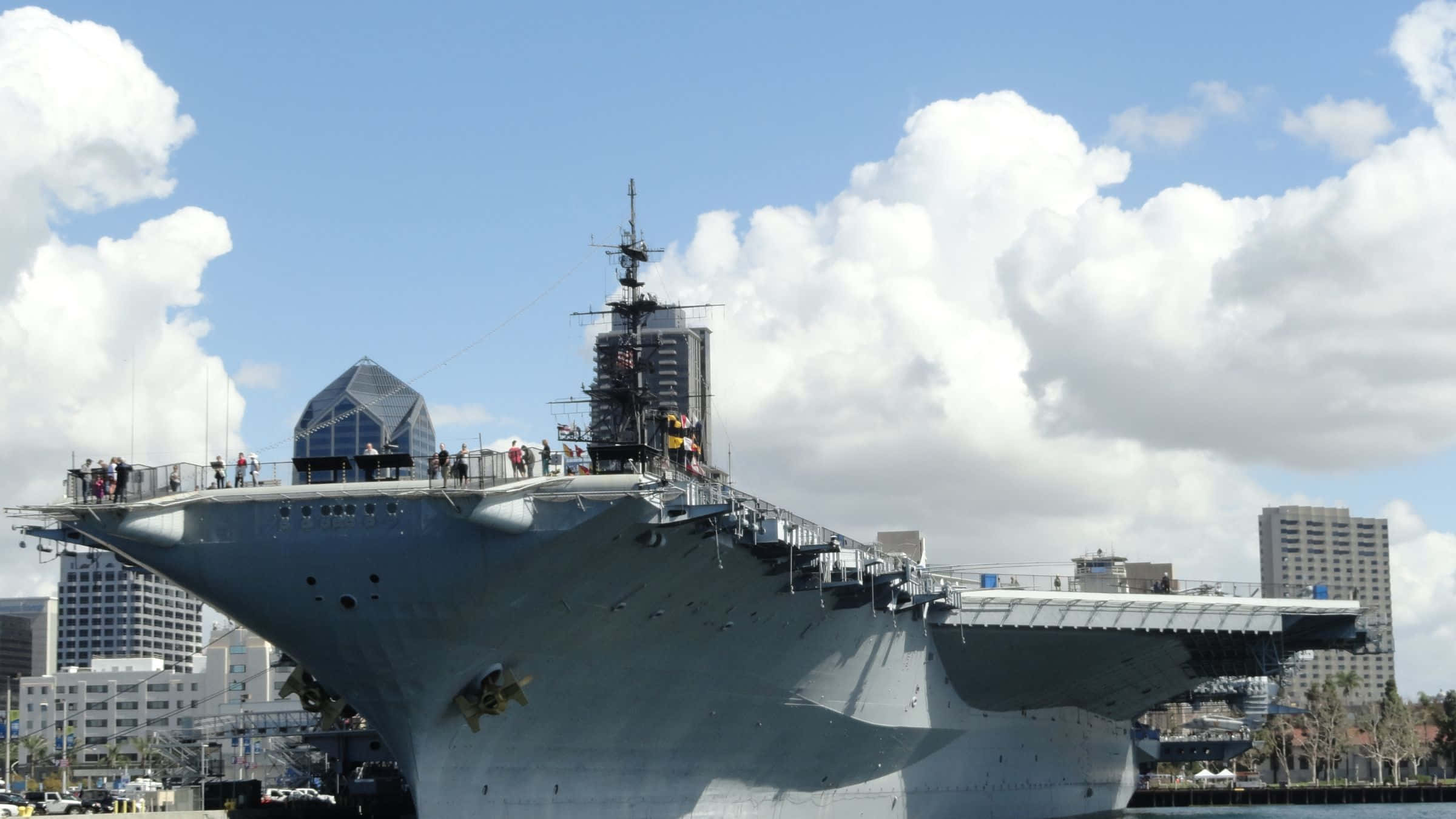 德州 桌布 壁紙 — 一艘巨大的灰色航空母艦停泊在港口，人們在甲板上行走，天空湛藍，白雲朵朵。