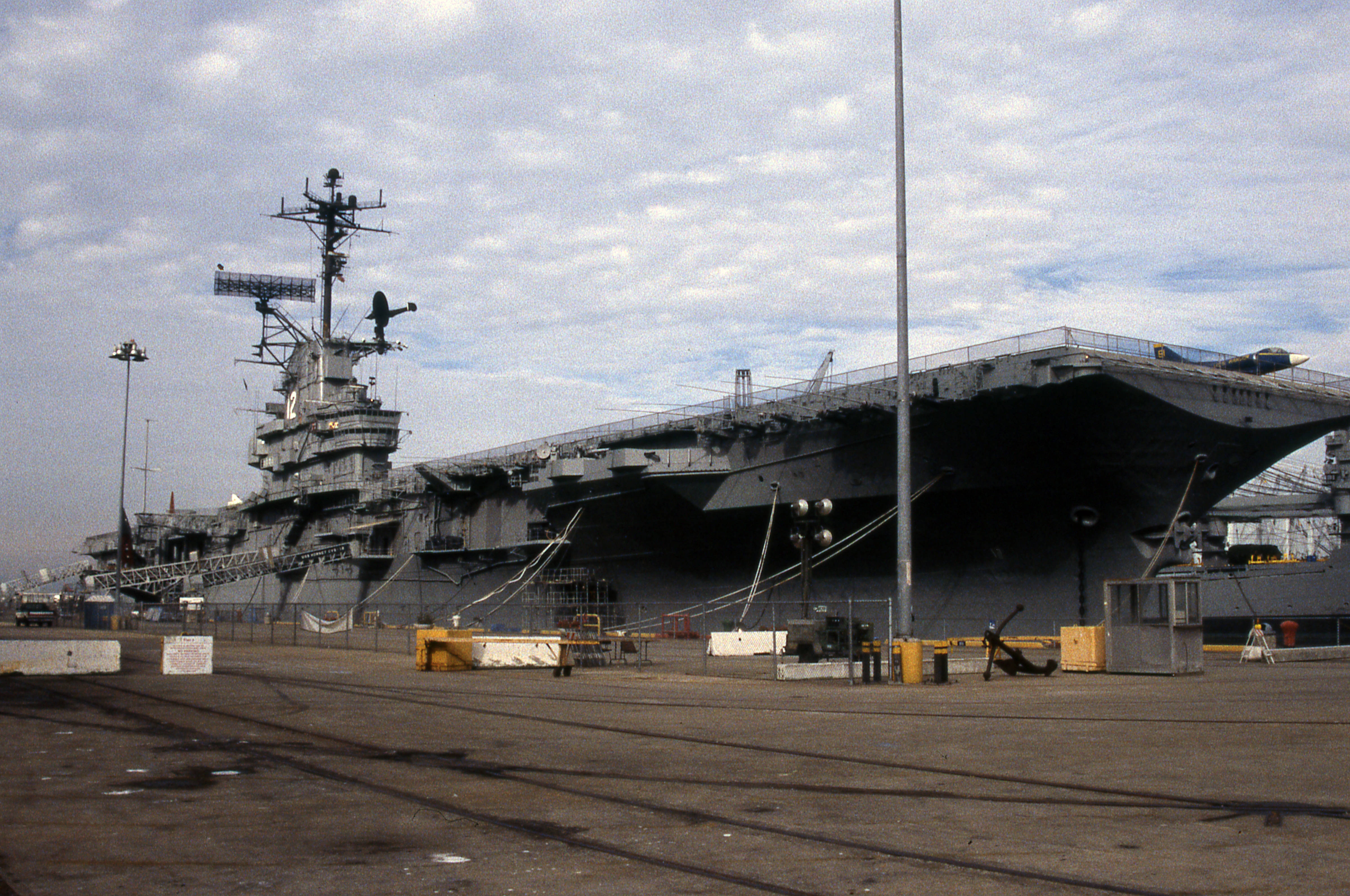 Warship 桌布 壁紙 — 一艘巨大的灰色航空母艦停泊在港口，天空多雲，附近有工業設備。