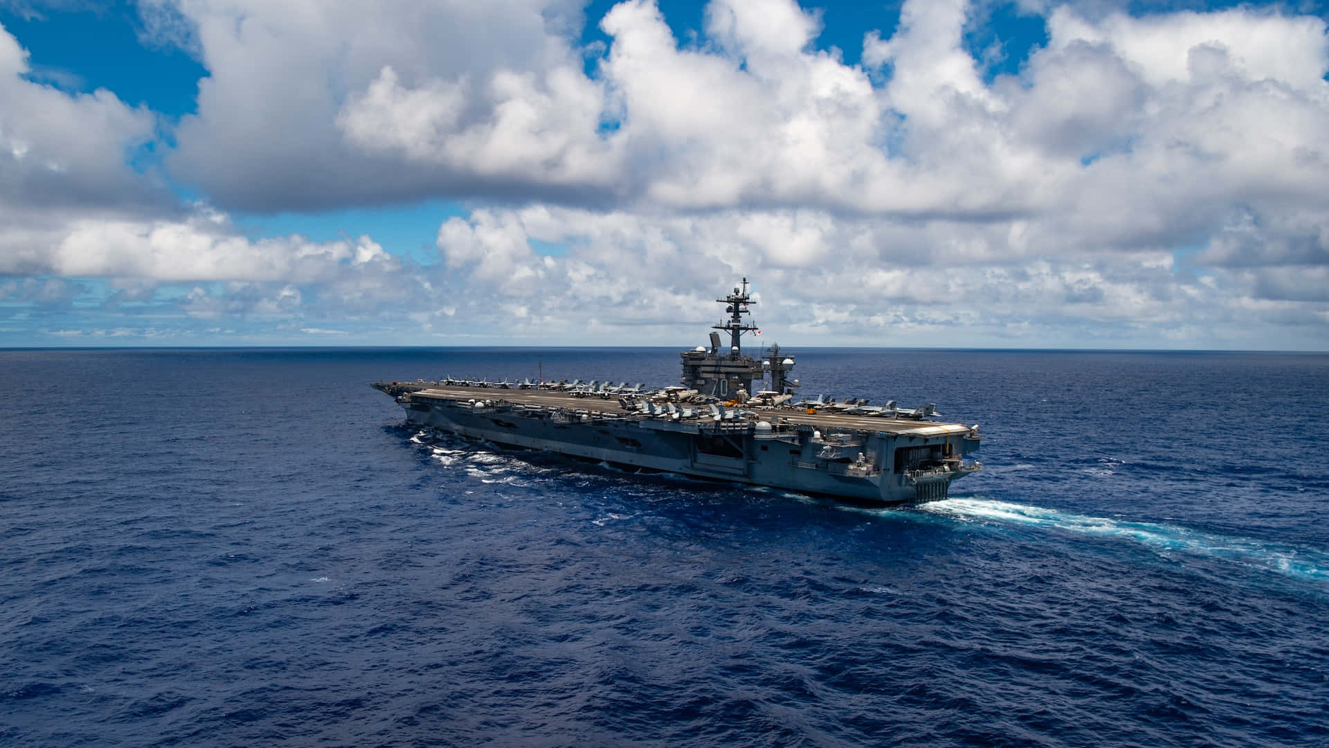 Navy 桌布 壁紙 — 一艘巨大的灰色航空母艦在晴朗的天空下，劃破深藍色的海浪，天空佈滿蓬鬆的白雲。