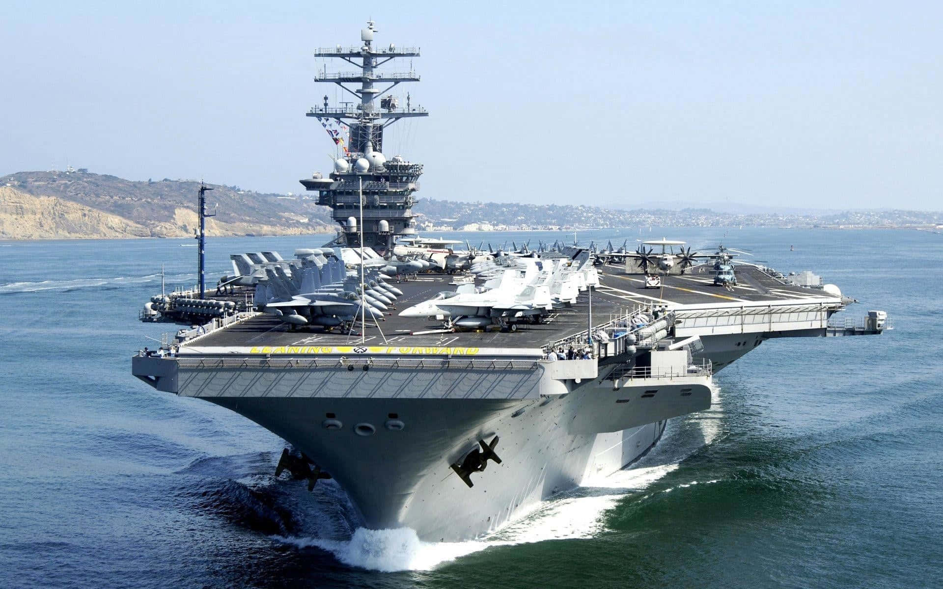 Navy 桌布 壁紙 — 一艘巨大的灰色航空母艦在藍色海洋中航行，甲板上排列著戰鬥機，天空晴朗。