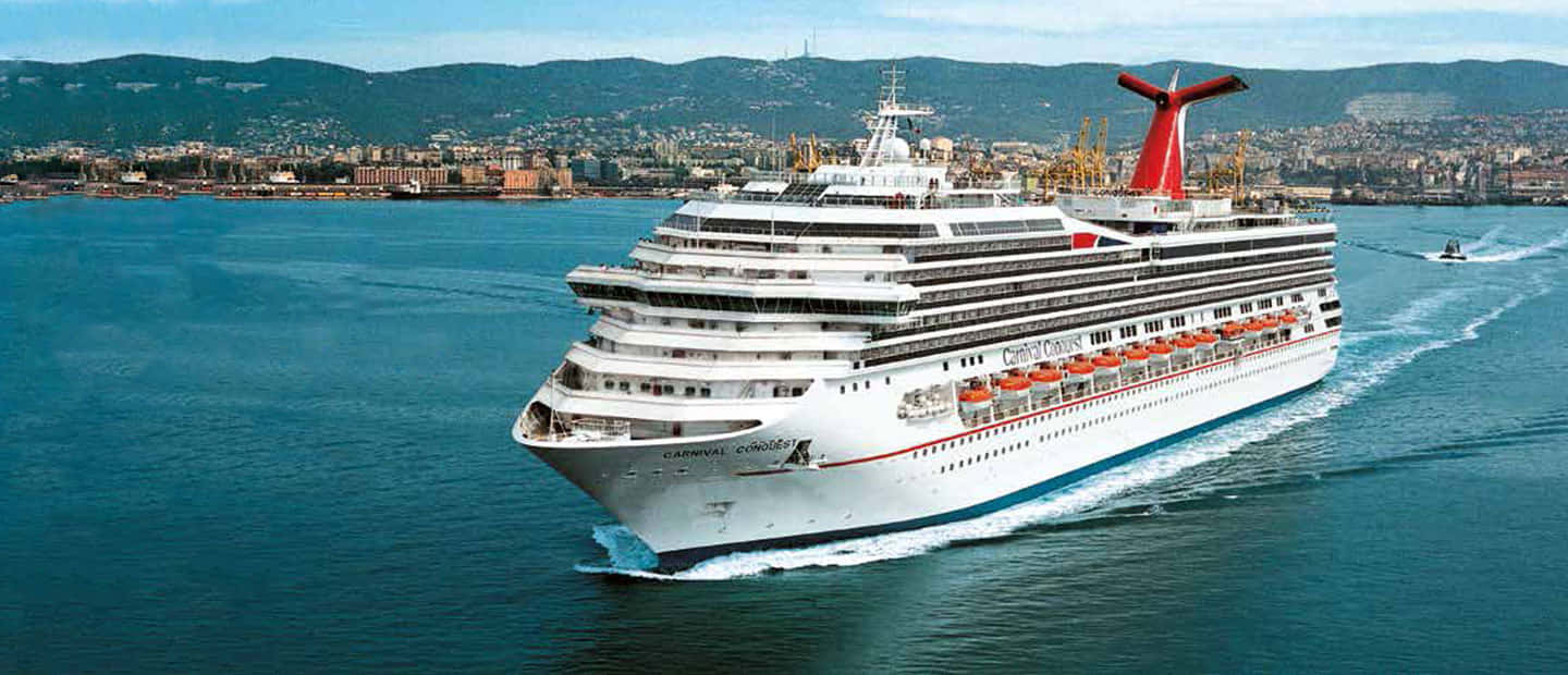 Carnival Conquest 桌布 壁紙 — 一艘巨大的白色嘉年華郵輪，帶有紅色煙囪，在深藍色水域中航行，靠近一個背景有綠色山丘的沿海城市。