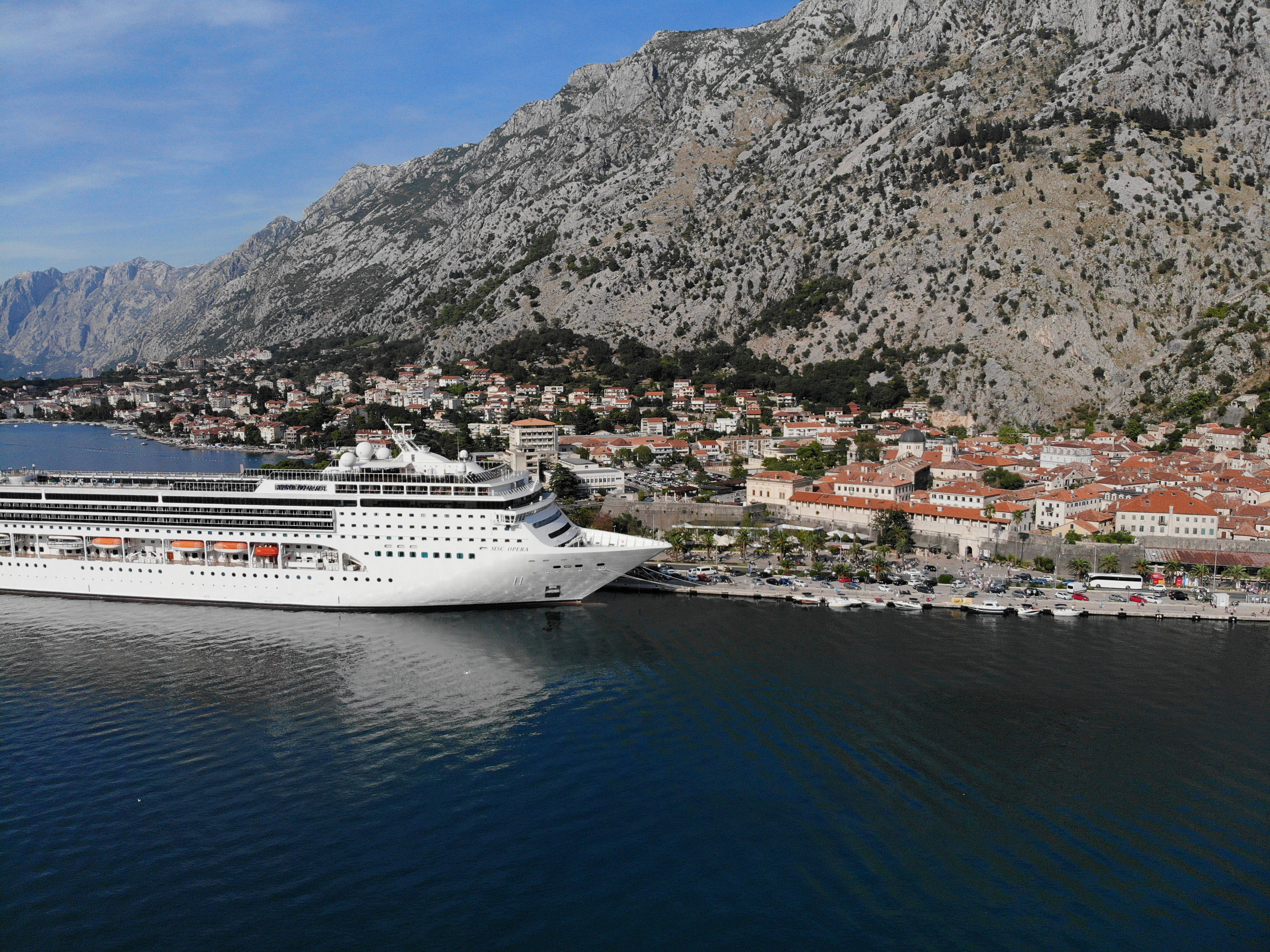 Montenegro 桌布 壁紙 — 一艘巨大的白色郵輪停泊在一個歷史悠久的沿海小鎮旁，小鎮擁有陶土色屋頂，依偎在 towering 岩石懸崖下，天空清澈湛藍。