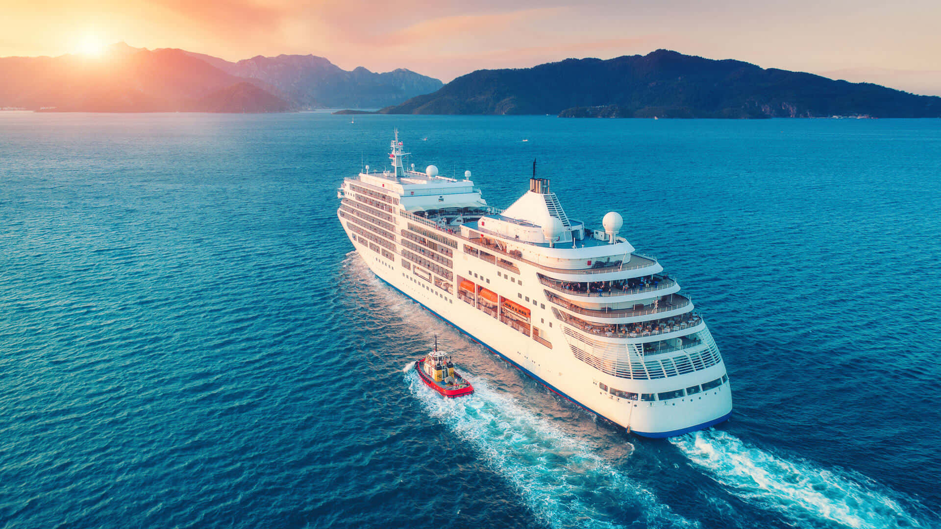 Cruise Ship 桌布 壁紙 — 一艘巨大的白色郵輪在日落時分航行於深藍色水域，背景有山脈，附近有一艘小型紅色拖船。