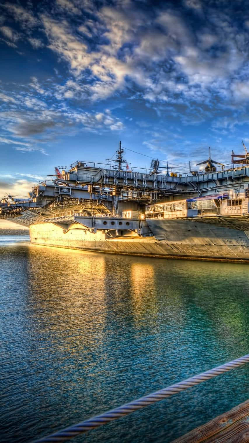 桌布 壁紙 — 一艘巨大的航空母艦停泊在平靜的水域，天空呈現戲劇性的藍色，金色的日落倒影在水面上閃爍。