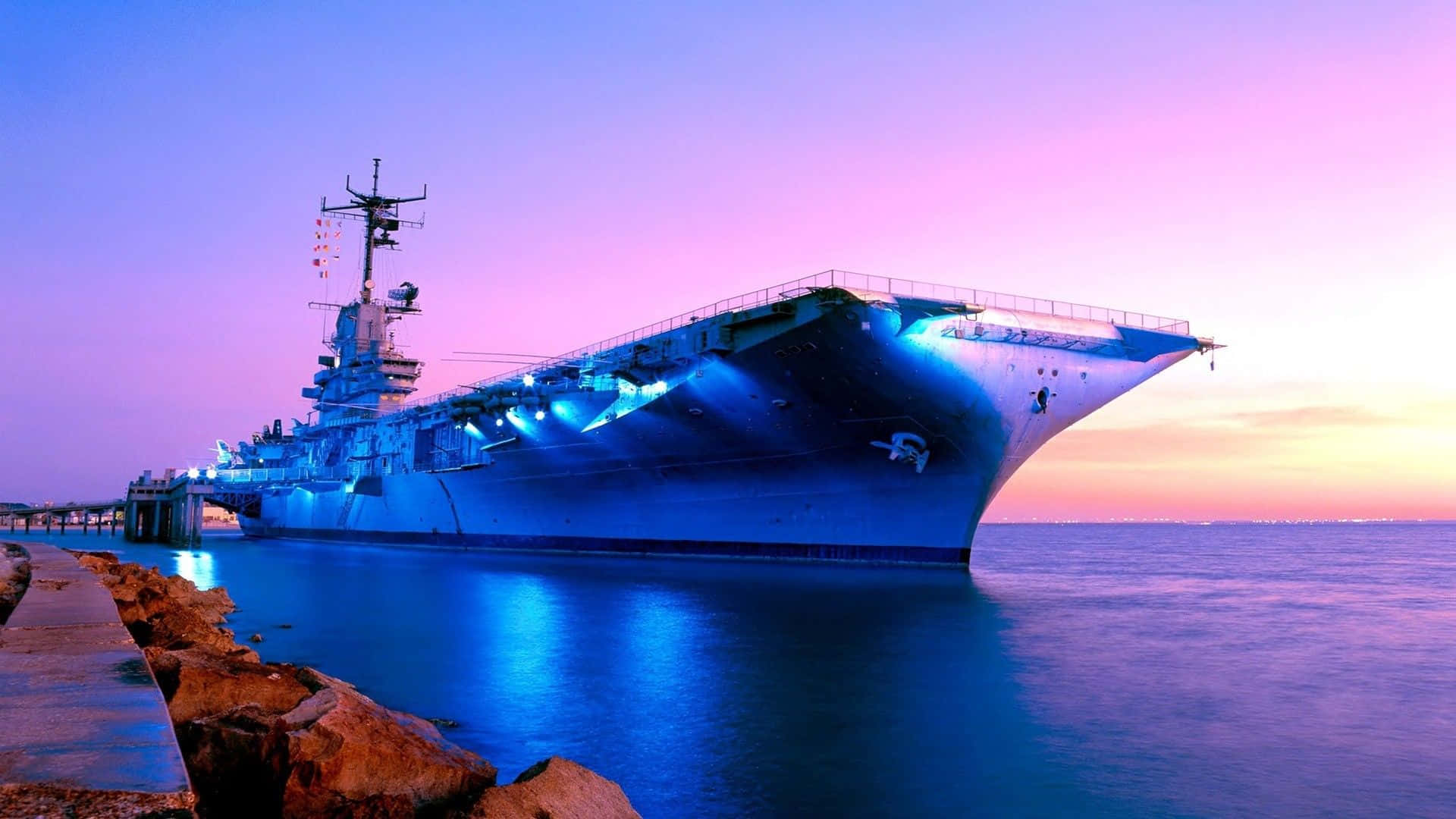 Navy 桌布 壁紙 — 一艘巨大的航空母艦在充滿活力的紫色和橙色日落天空下發出藍色光芒。