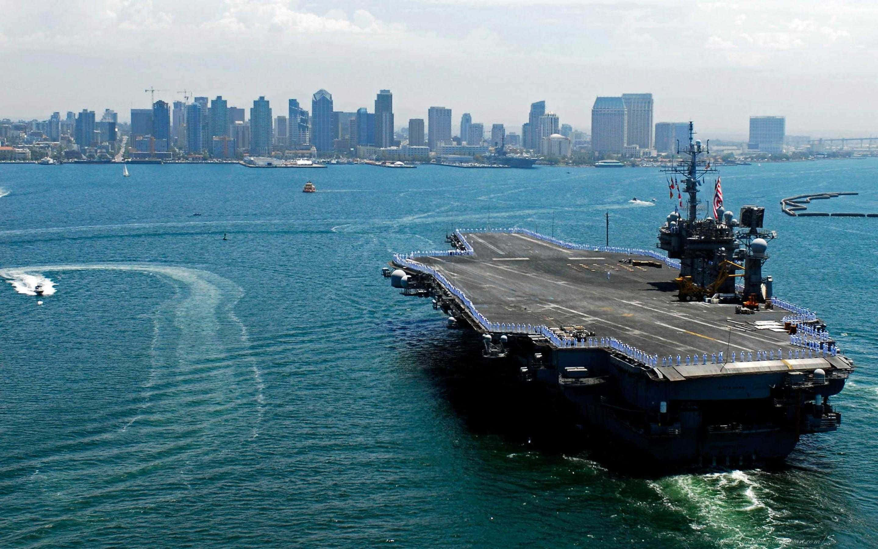 Navy 桌布 壁紙 — 一艘巨大的航空母艦在碧綠色的水域中航行，背景是晴朗天空下現代城市的天際線。