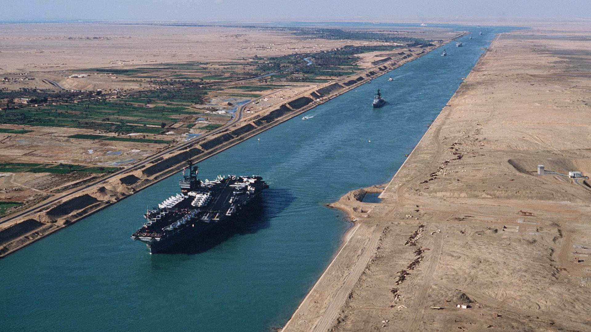 Cargo Ship 桌布 壁紙 — 一艘巨大的航空母艦在蘇伊士運河航行，兩側是乾旱的沙漠和綠色的農業地帶，天空晴朗。