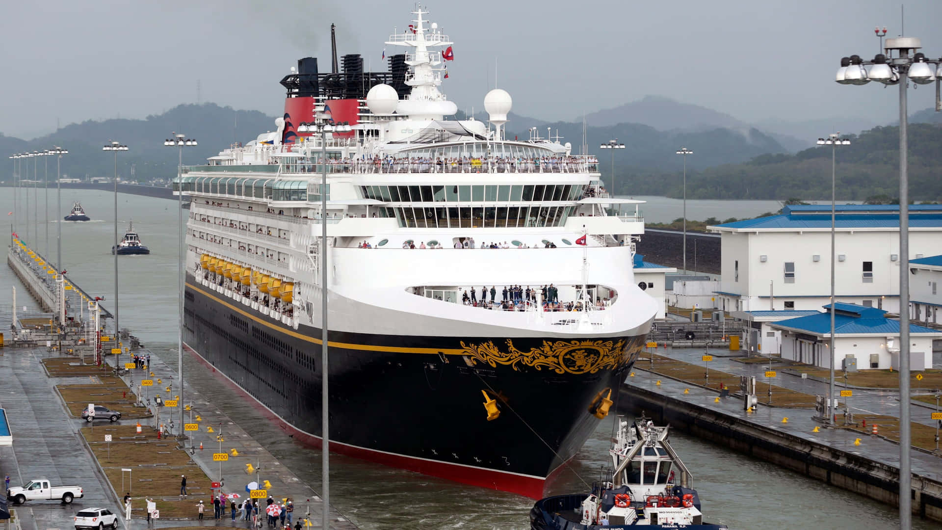 shipping 桌布 壁紙 — 一艘巨大的迪士尼郵輪航行通過巴拿馬運河，背景有山脈，碼頭上有民眾觀看。