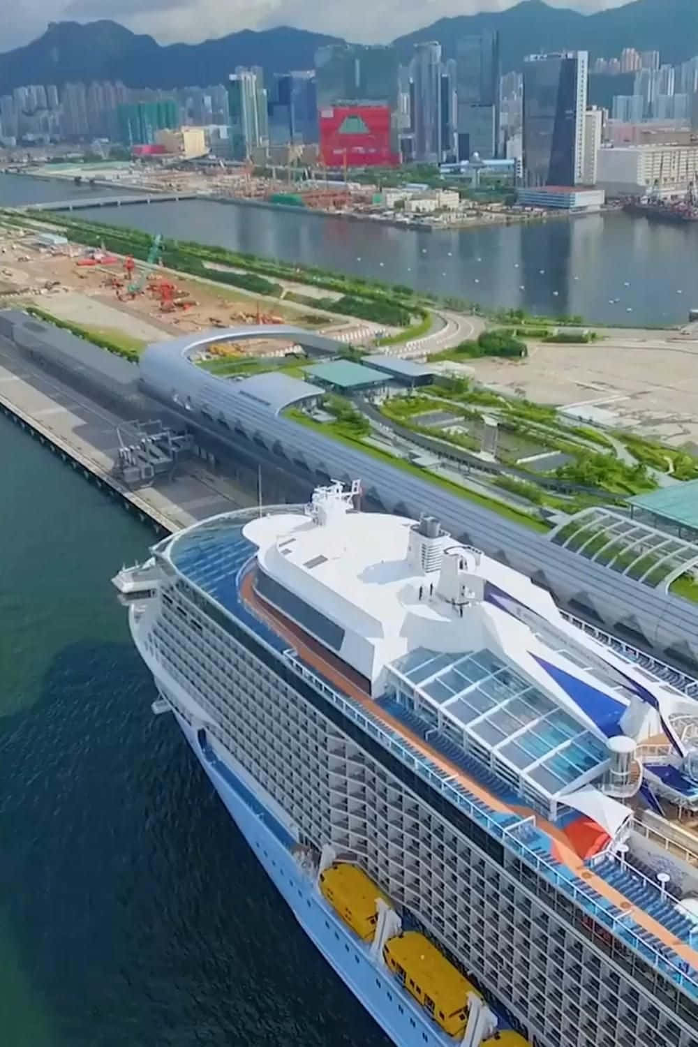 Cruise 桌布 壁紙 — 一艘巨大的郵輪停靠在現代化的港口，背景是高聳的城市摩天大樓和鬱鬱蔥蔥的綠色山脈，天空晴朗。