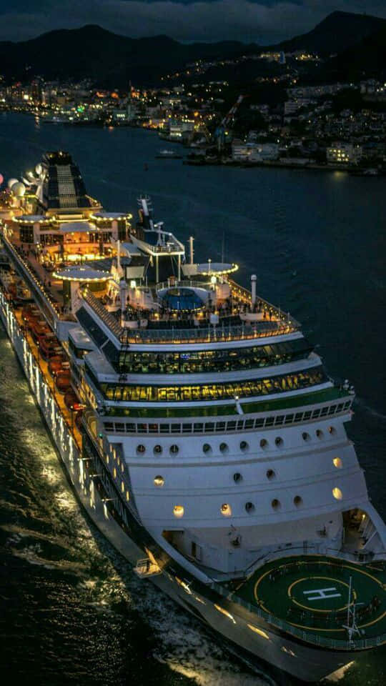 Cruise 桌布 壁紙 — 一艘巨大的郵輪在夜間駛過黑暗的港口，發出溫暖的光芒，背景是城市景觀和山脈。