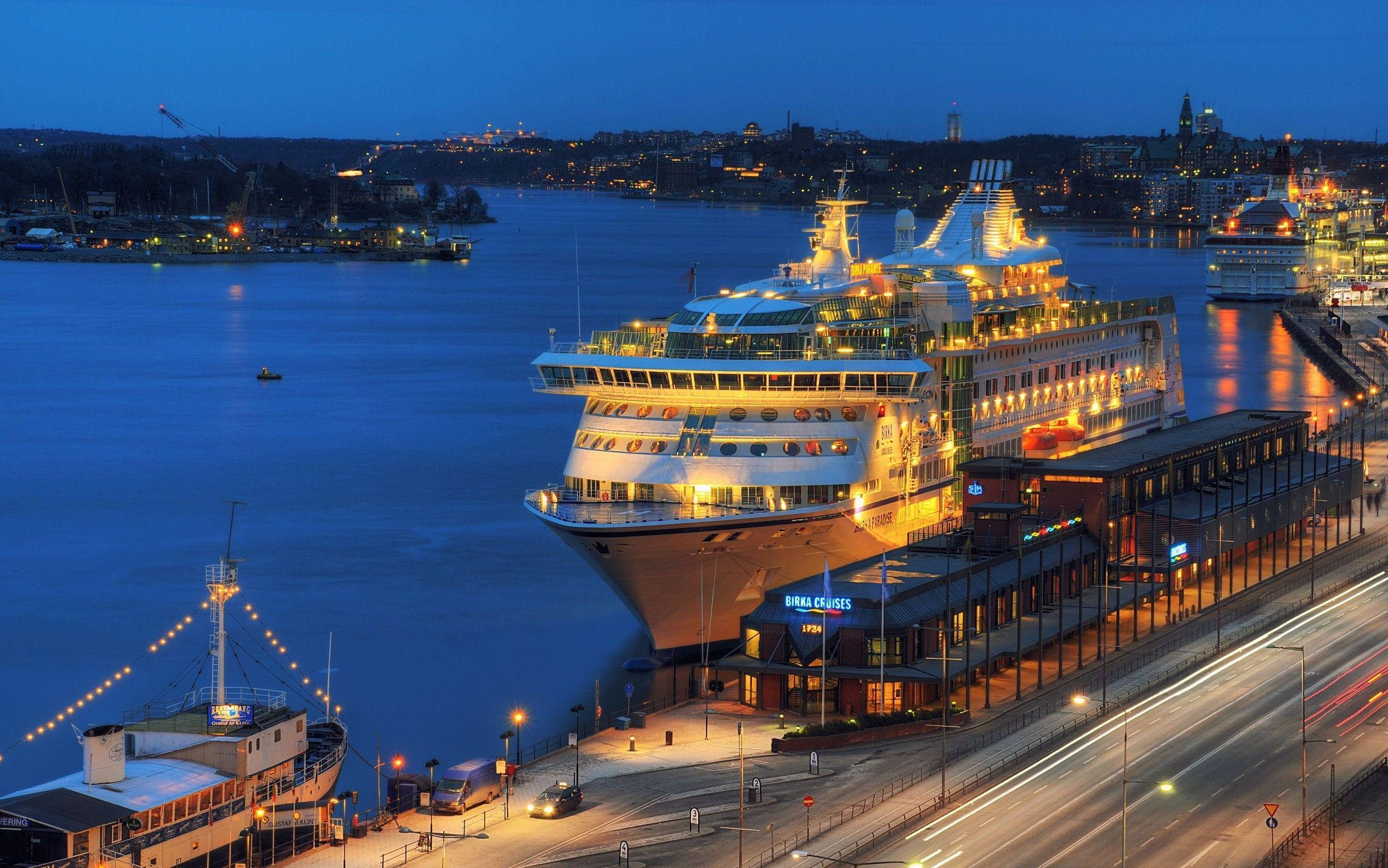 Cruise Ship 桌布 壁紙 — 一艘巨大的郵輪在黃昏時分溫暖地發光，停靠在繁忙的港口，城市燈光倒映在平靜的水面上。