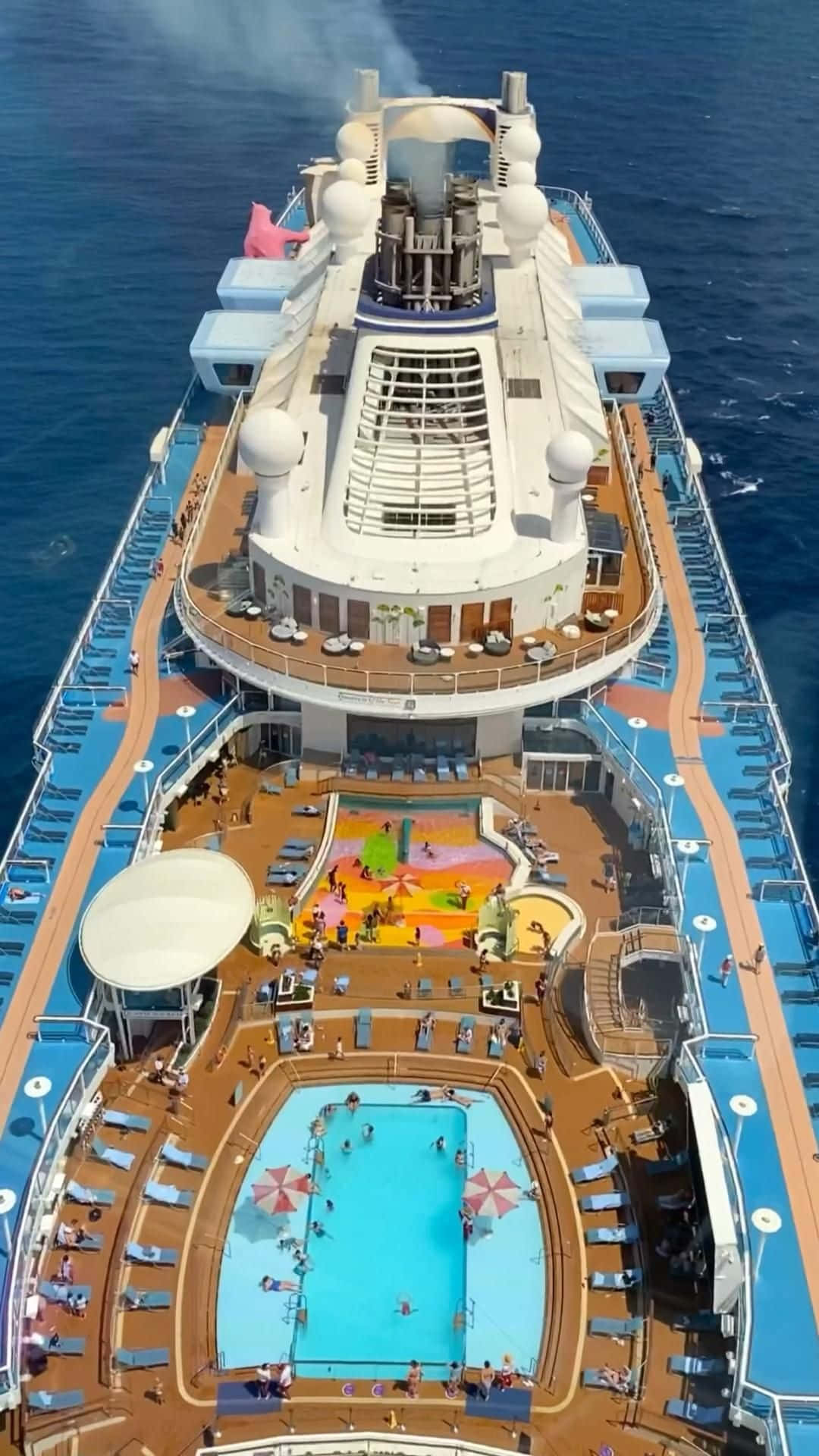 Cruise 桌布 壁紙 — 一艘巨大郵輪頂層甲板的鳥瞰圖，設有游泳池、躺椅和色彩繽紛的水滑梯，背景是深藍色的海洋。
