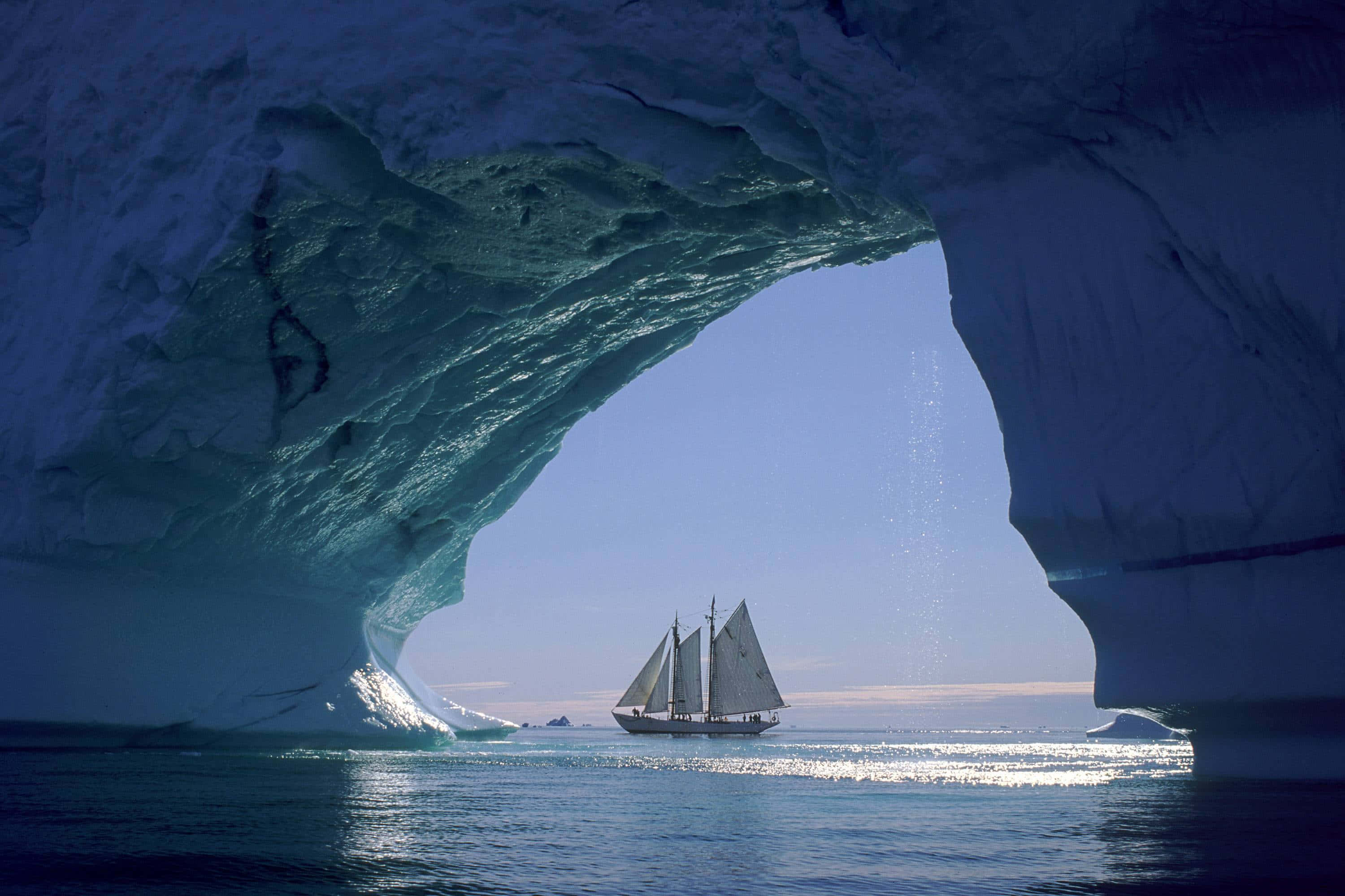 高清 桌布 壁紙 — 一艘帆船在南極巨大的冰拱門下滑行，陽光穿過藍色的冰層。