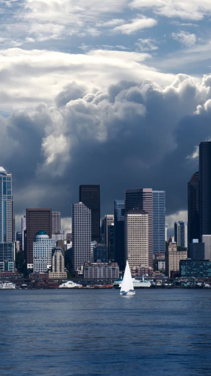 Skyscraper 桌布 壁紙 — 一艘帆船在平靜的水面上滑行，上方是現代城市天際線上空翻湧的戲劇性雲層。